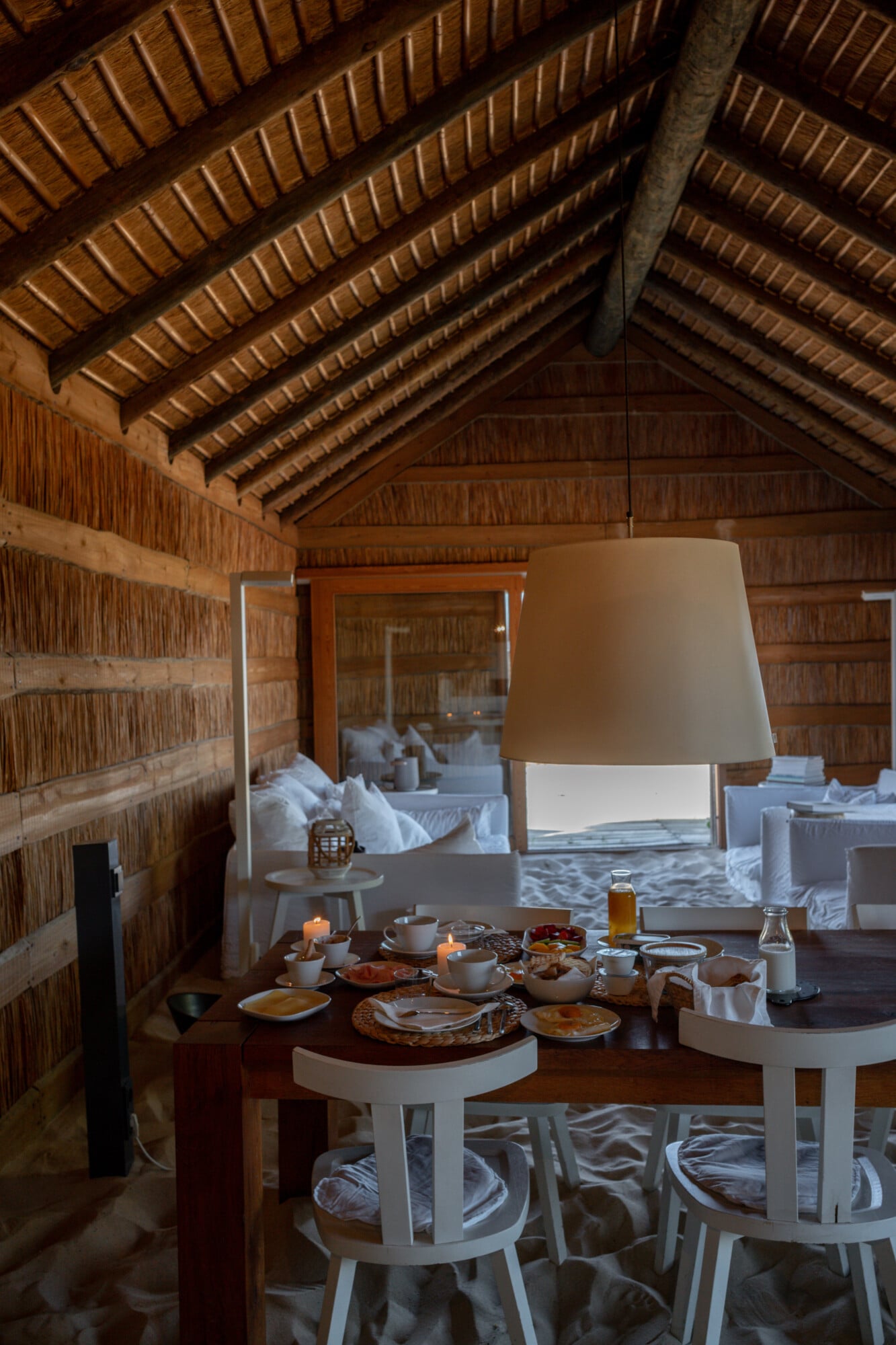 Casas na Areia, Europe — Dining pavilion with exposed brick walls, wooden beam ceiling, and pendant lamp overlooking ocean view through glass doors.