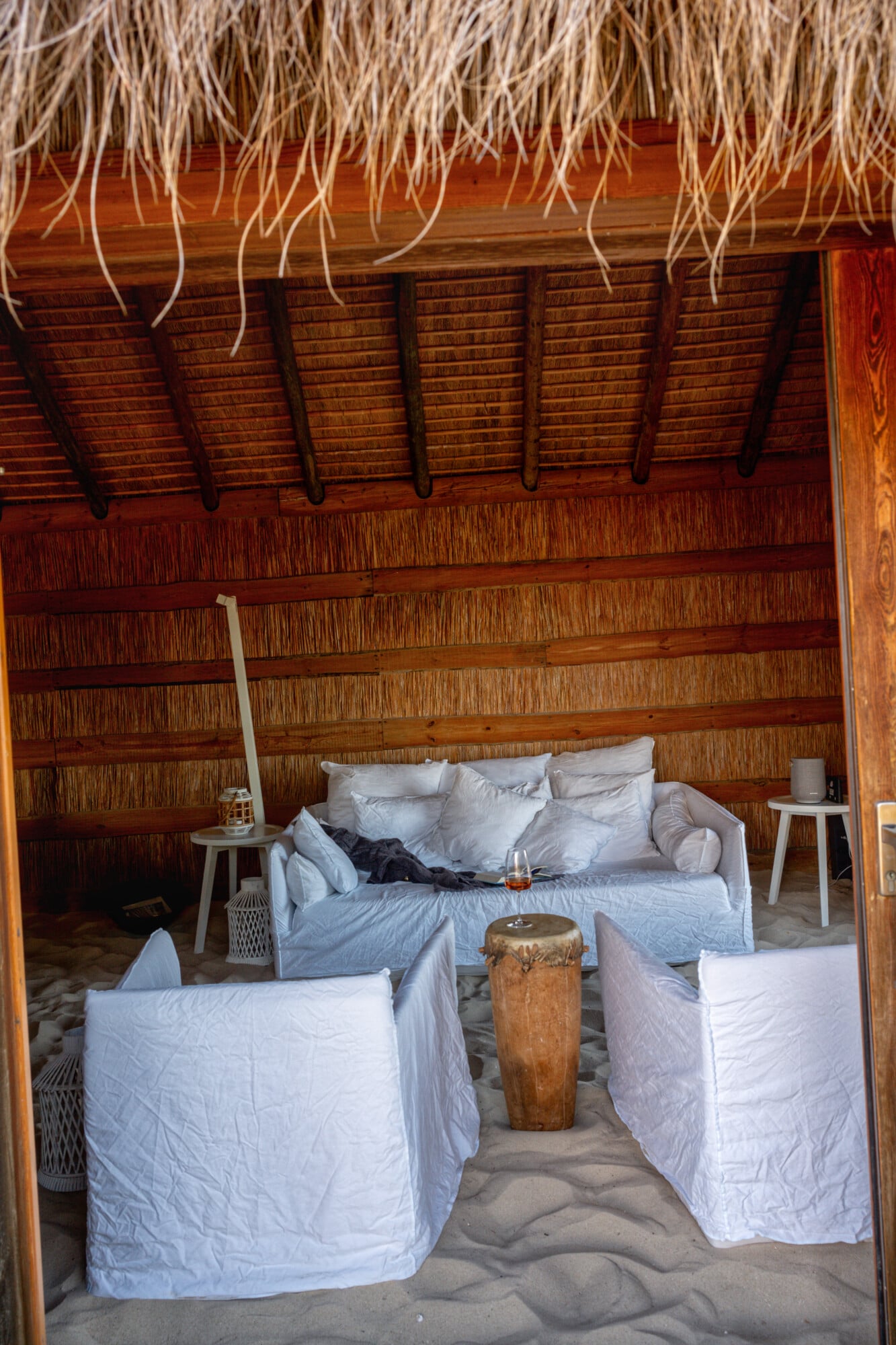 Casas na Areia, Europe — Thatched-roof beach pavilion with white linen seating, wooden walls, sand floors, and carved drum accent table.
