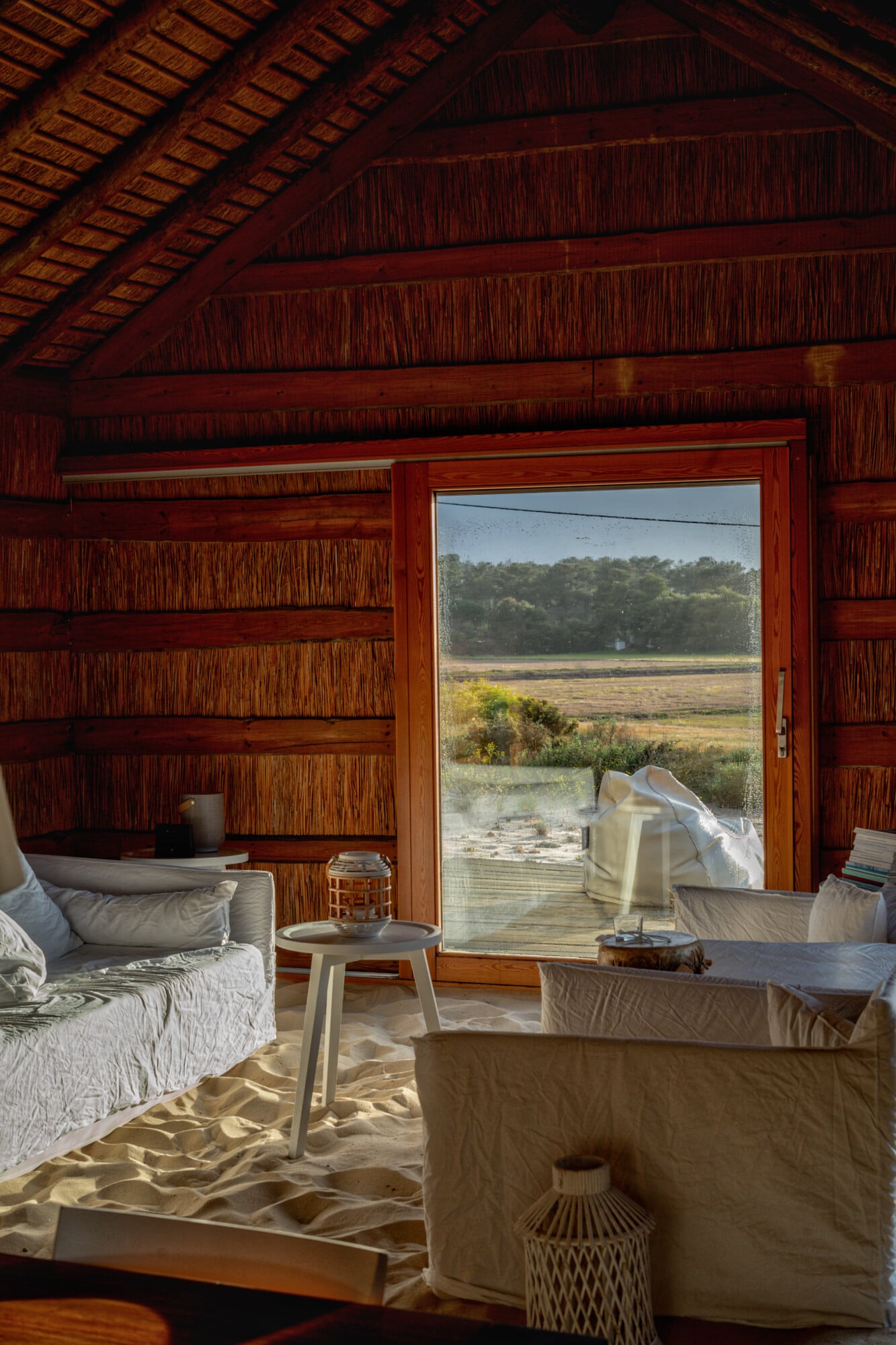 Casas na Areia, Europe — Wooden interior with rattan-clad ceiling, sand floor, linen furnishings, and open glass doors overlooking marshland and trees.