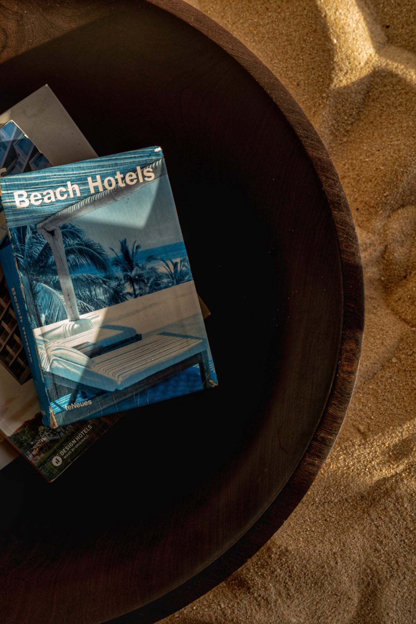 Casas na Areia, Europe — Beach Hotels design book on dark wood table with warm sand-colored textiles and dappled sunlight, coastal hospitality aesthetic.