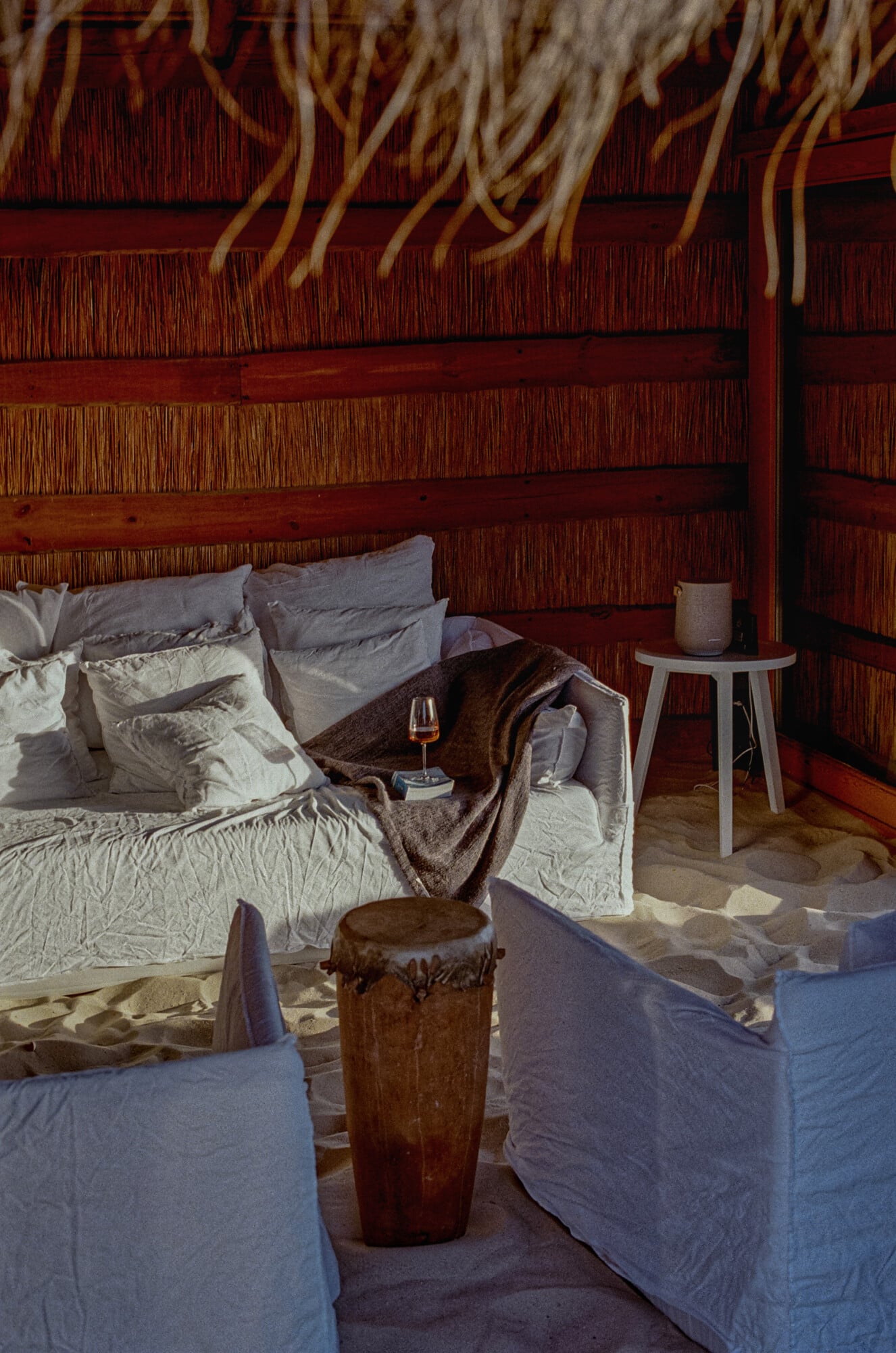 Casas na Areia, Europe — Coastal bedroom with woven rattan headboard, linen bedding, wooden drum stool, and dappled sunlight on sand floor.