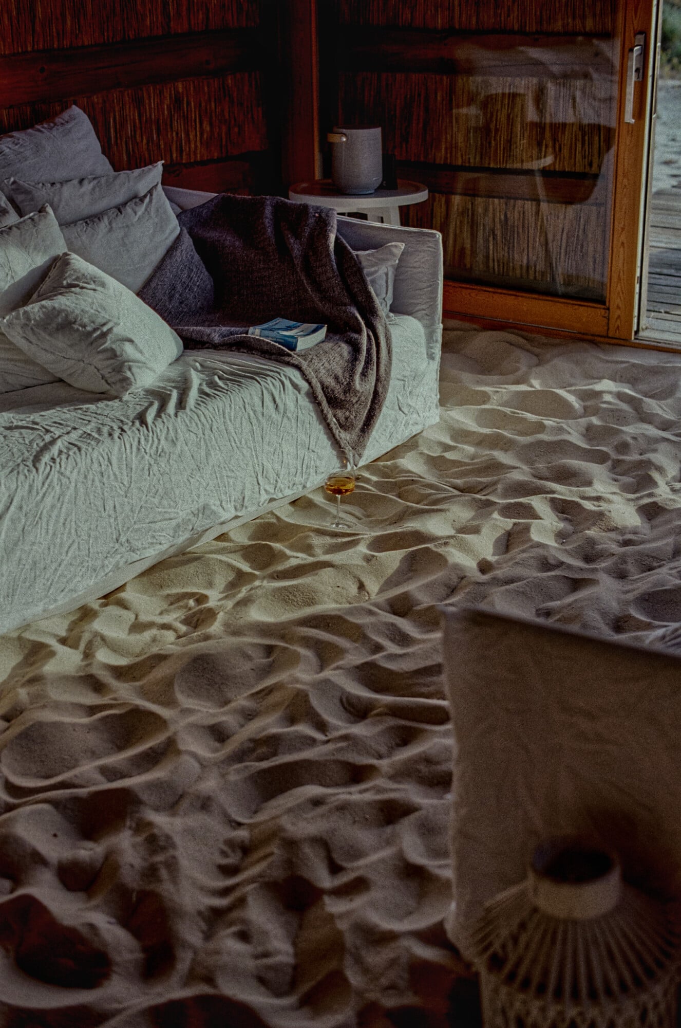 Casas na Areia, Europe — Bedroom with sand floor, linen sofa, dark wooden storage, natural light, minimalist coastal interior design.