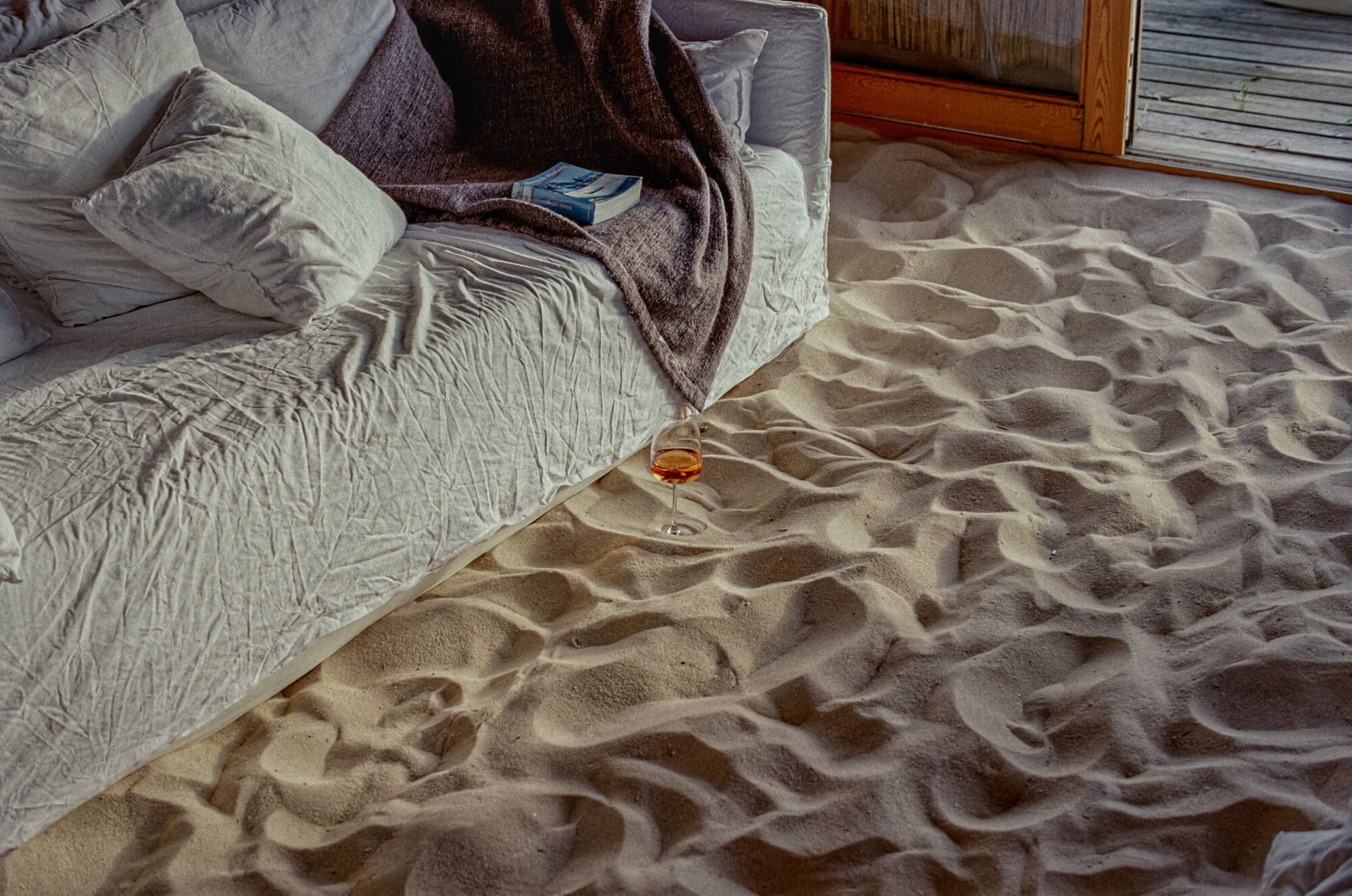 A glass of amber liquor rests on textured sand beside a bed with rumpled linen sheets and a dark throw blanket.
