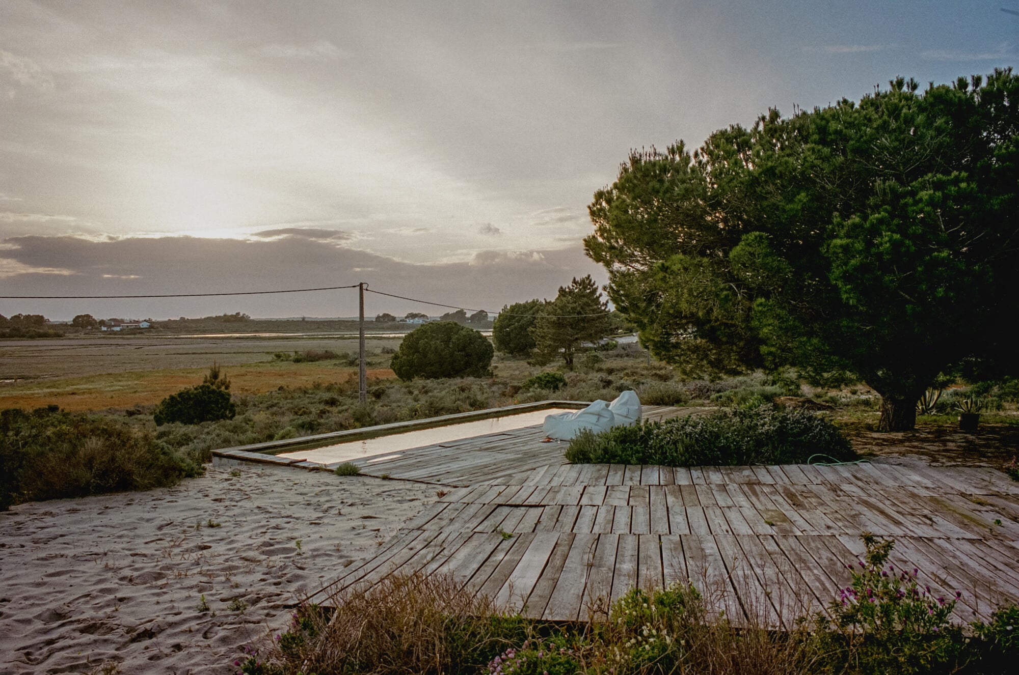 Casas na Areia, Europe — Weathered wooden deck overlooking salt marshes at dusk, with minimalist pool structure and pine trees under clouded sky.