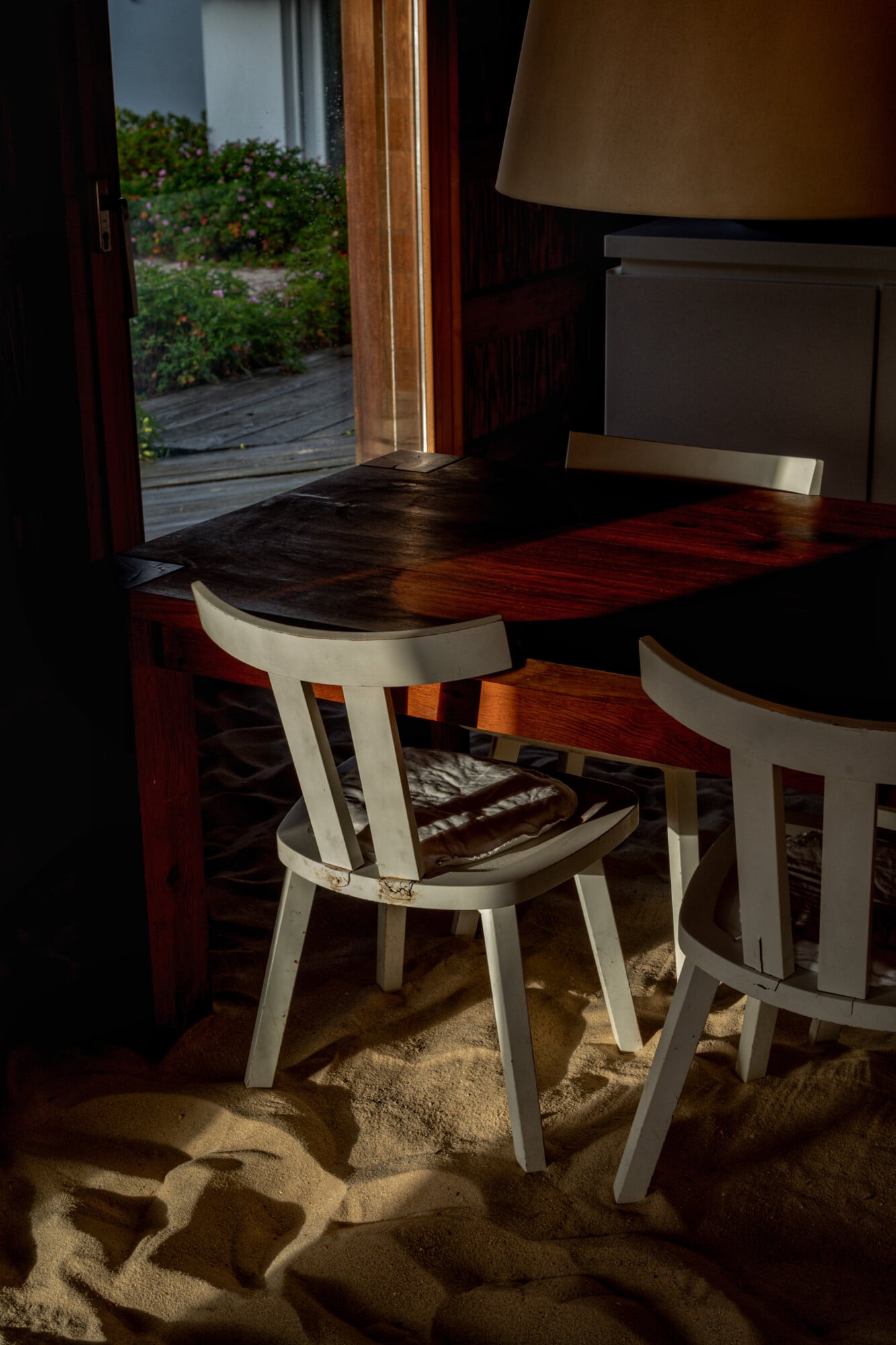 Casas na Areia, Europe — White bentwood chairs at timber desk with sand floor, warm sunlight streaming through open doors onto coastal garden beyond.