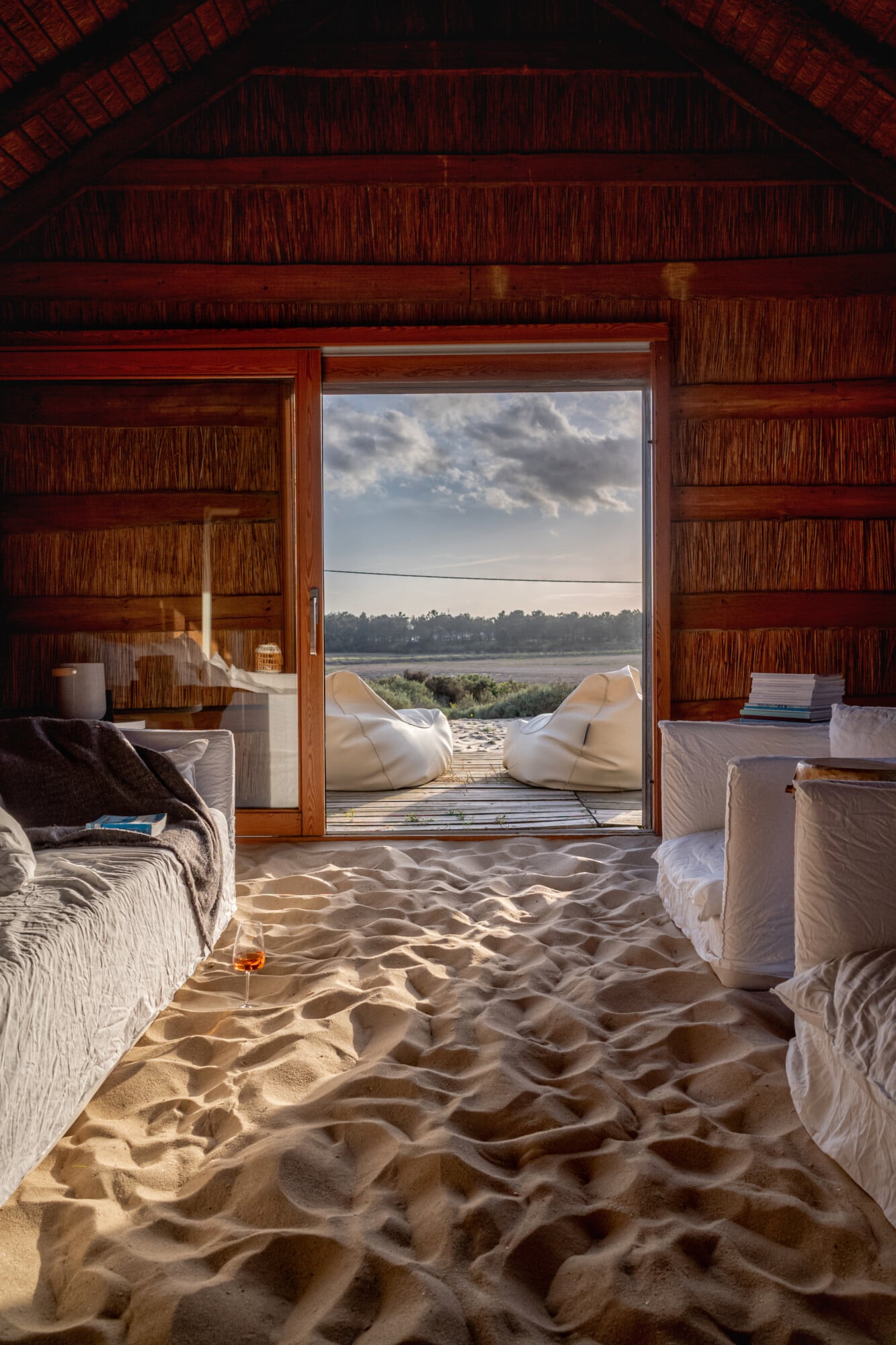 Casas na Areia, Europe — Wooden beach villa interior with sand flooring, white linen furnishings, and expansive water views through open glass doors.
