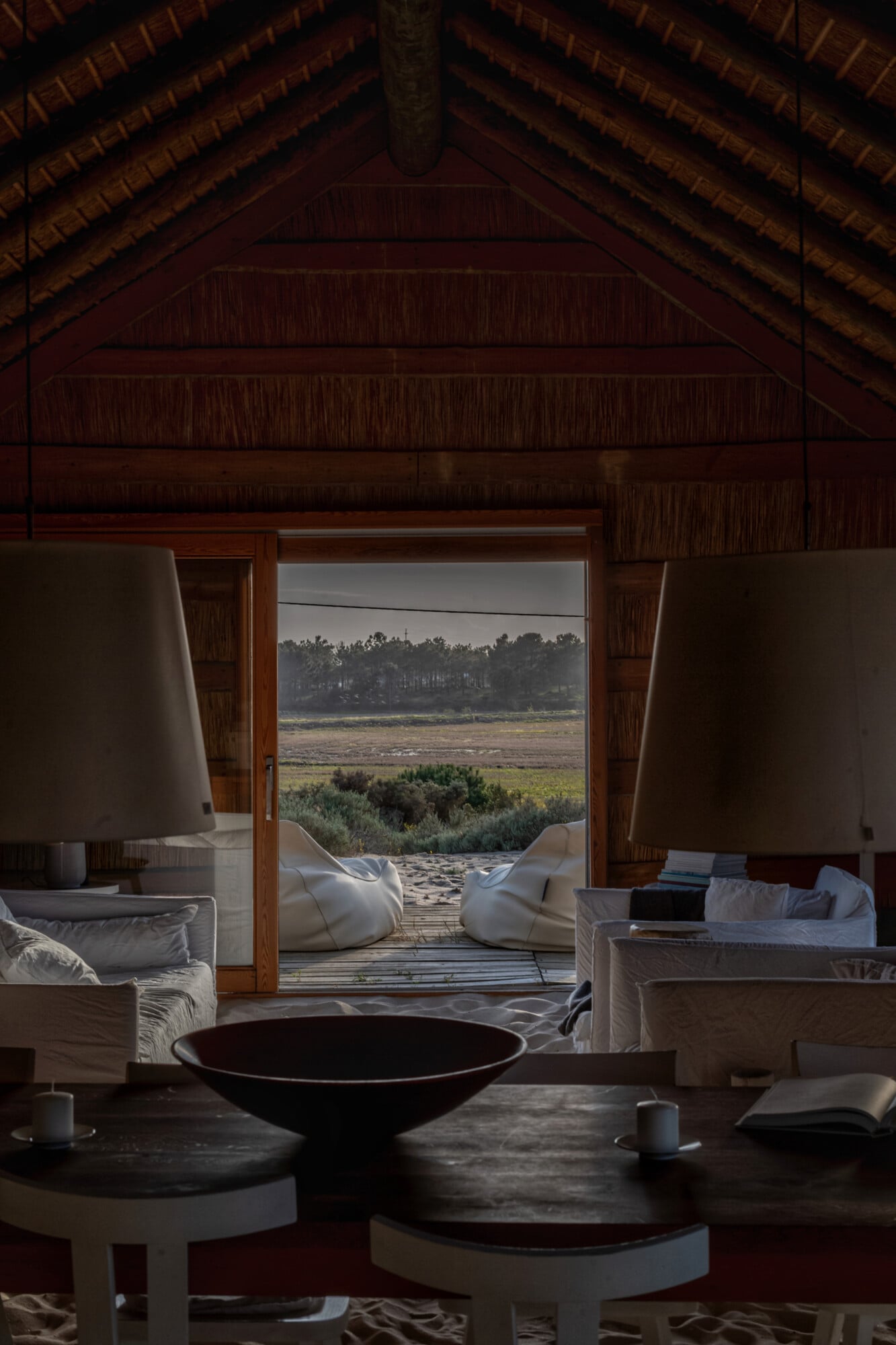 Casas na Areia, Europe — Open-plan pavilion with brick vaulted ceiling, teak doors framing wetland views, white sculptural seating, black ceramic vessel, natural light.