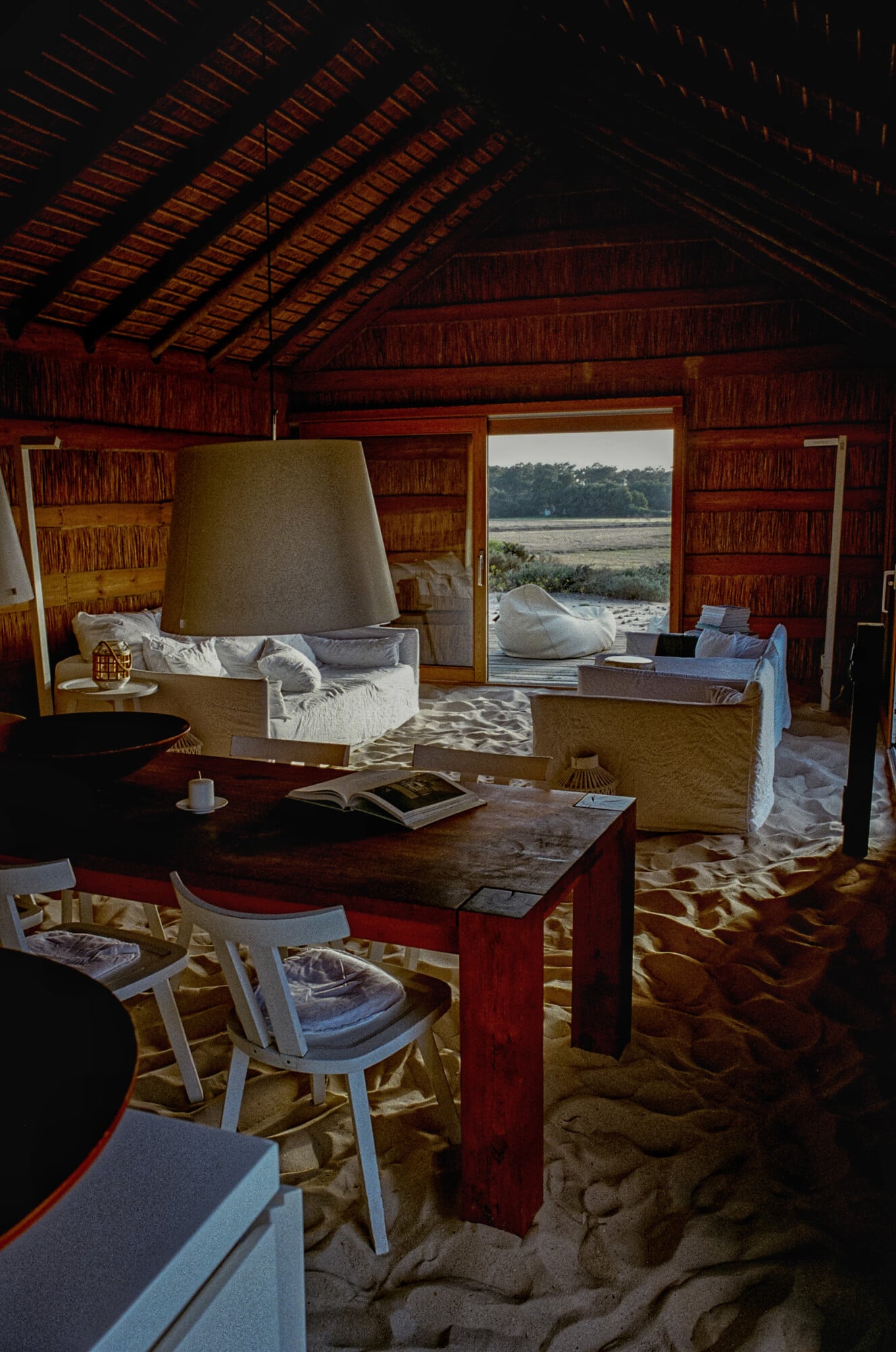 Casas na Areia, Europe — Coastal villa interior with reddish timber walls, sand flooring, cream furniture, and waterfront views through open doors.