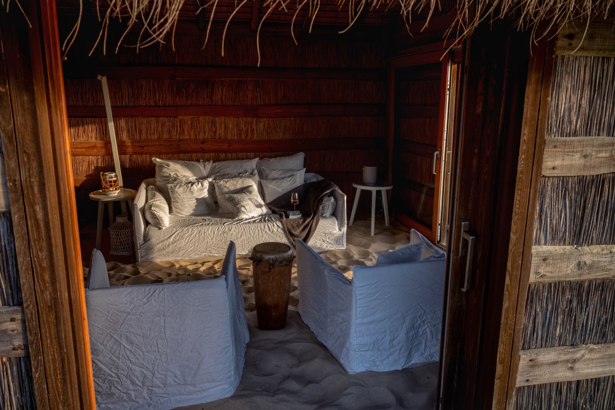 Casas na Areia, Europe — Candlelit bedroom pavilion with woven palm walls, linen-draped furniture, and sand flooring creating intimate tropical ambiance.