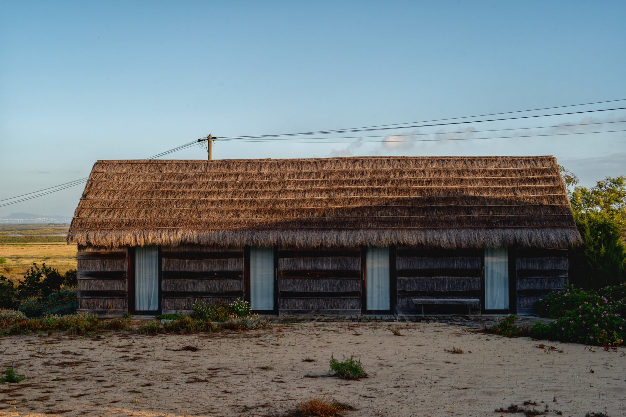 Casas na Areia, Europe — Thatched-roof beach house with weathered timber walls, white-trimmed windows, and sand courtyard overlooking coastal landscape.