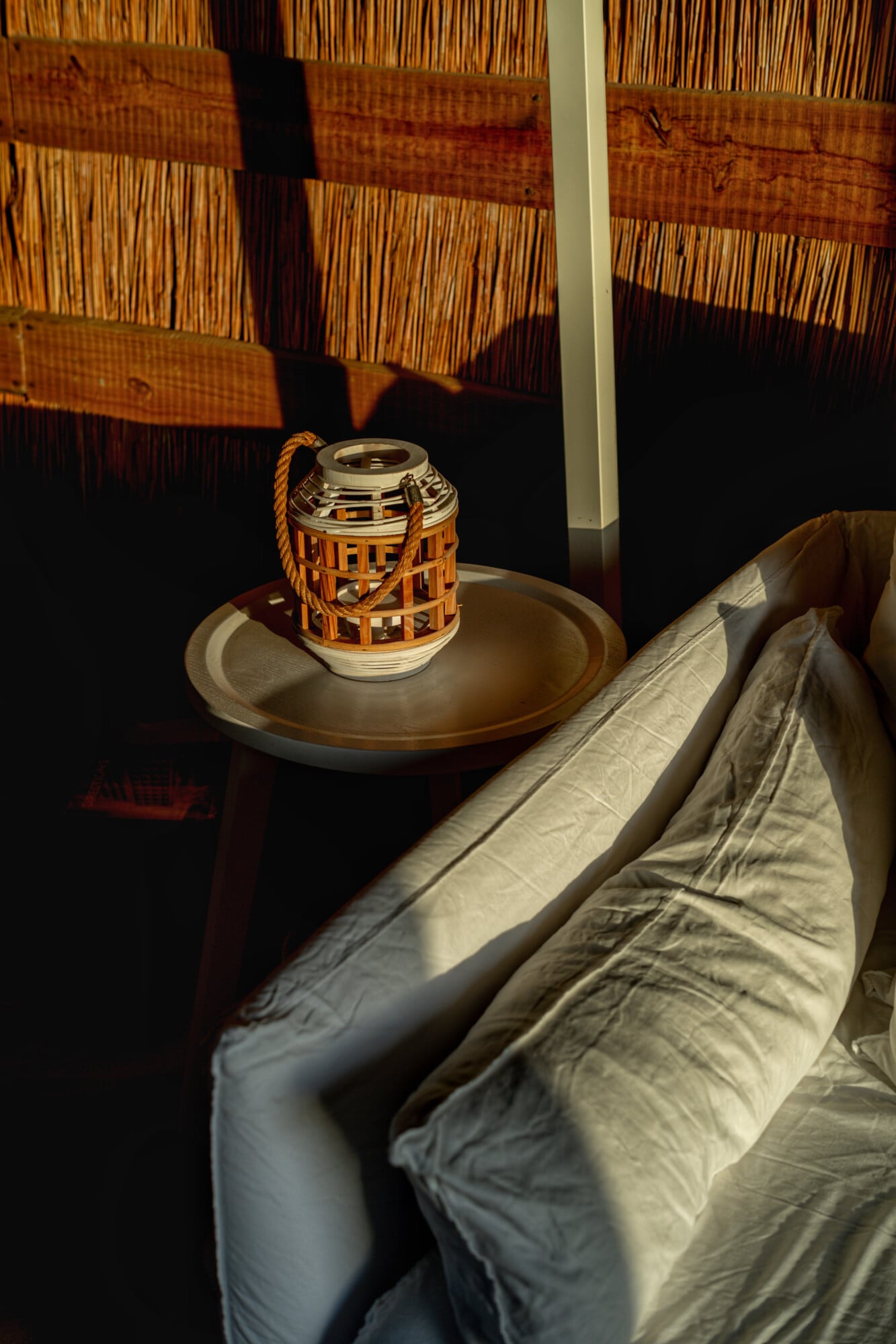 Casas na Areia, Europe — Woven wooden lantern with rope handle on ceramic dish beside linen bedding, afternoon light casting shadows on woven wall panels.