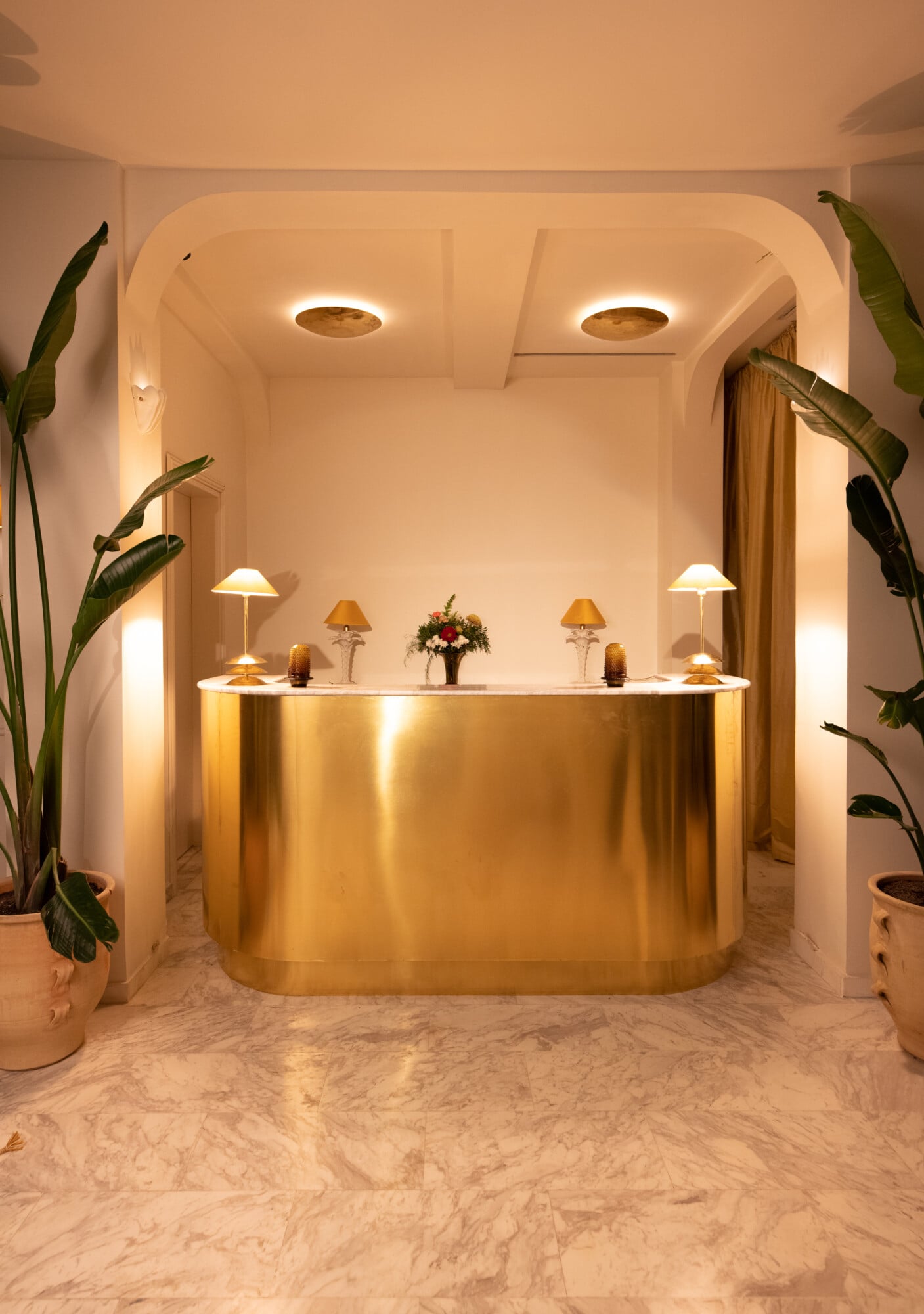 Apollo Palm, Europe — Brass curved reception desk with marble flooring, warm ambient lighting, potted plants, and minimalist luxury hospitality interior design.