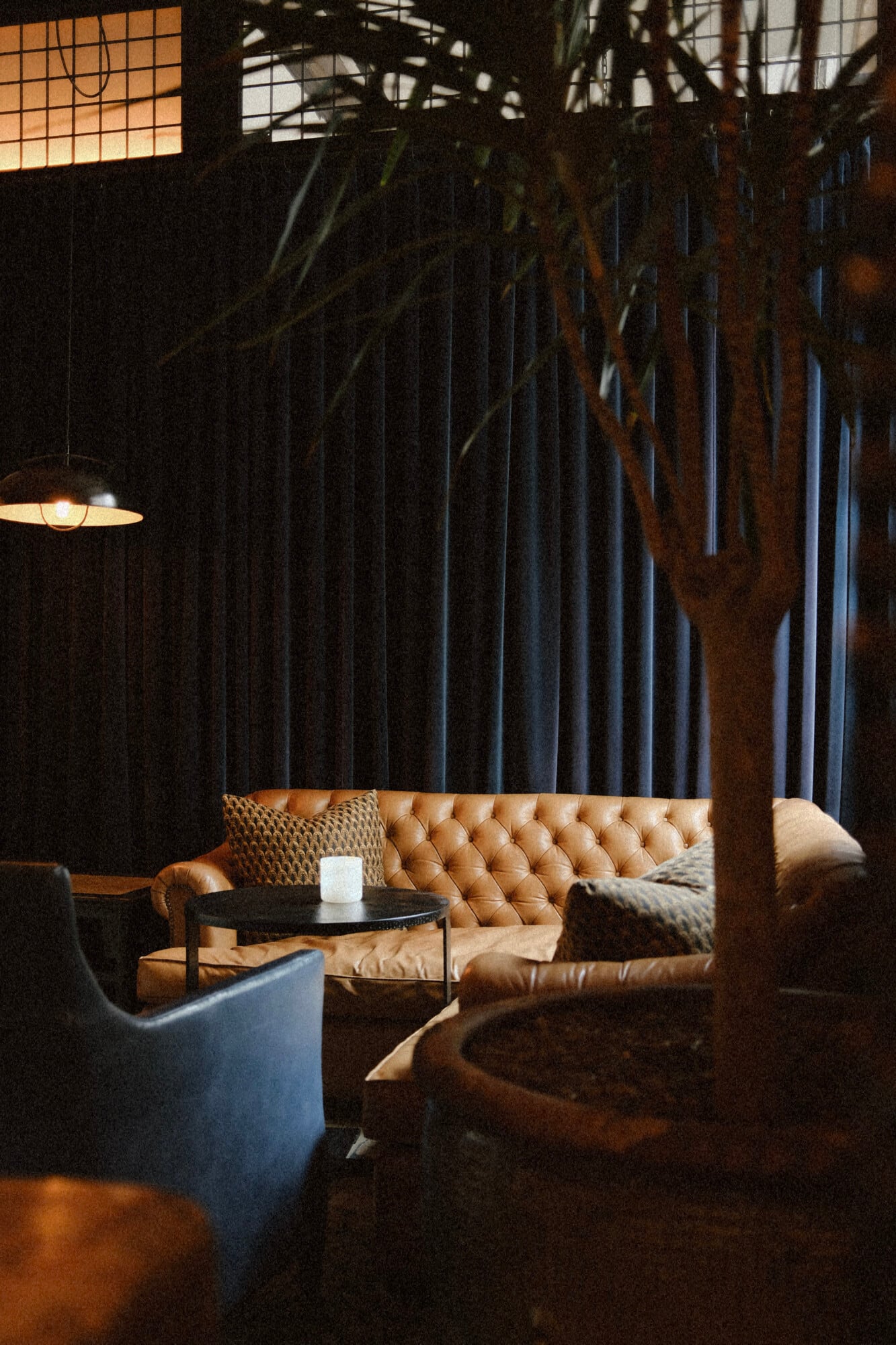 The Greenwich Hotel, North America — Tufted caramel leather banquette with black side table, pendant lighting, and industrial-chic exposed brick walls in moody lounge setting.
