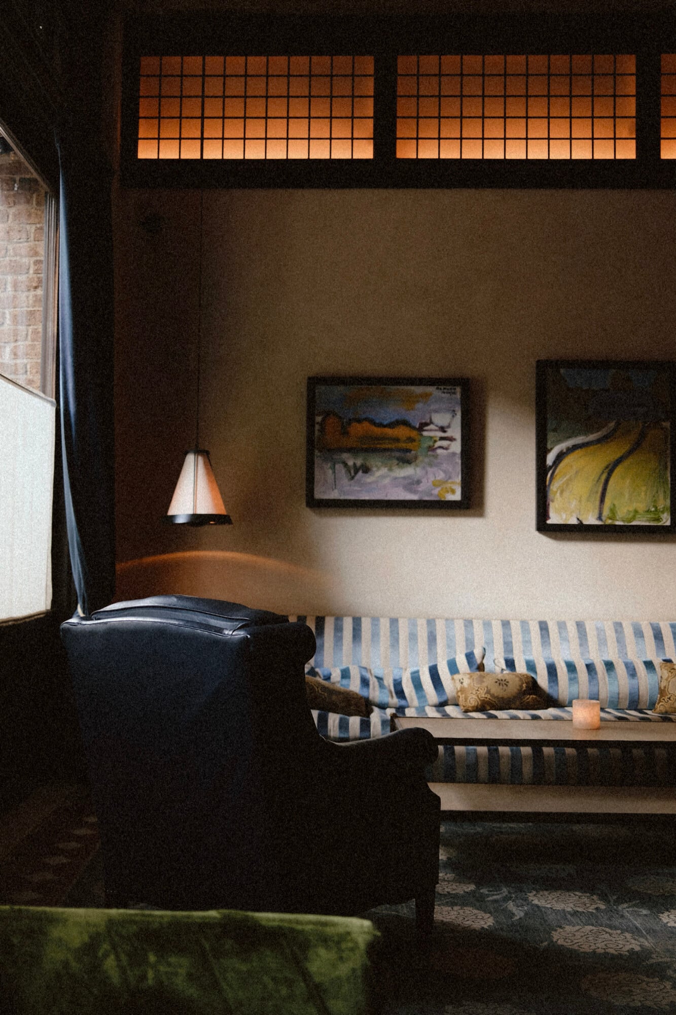 The Greenwich Hotel, New York, United States — Minimalist hotel room with striped daybed, pendant lamp, framed artwork, and illuminated transom windows in warm ambient light.