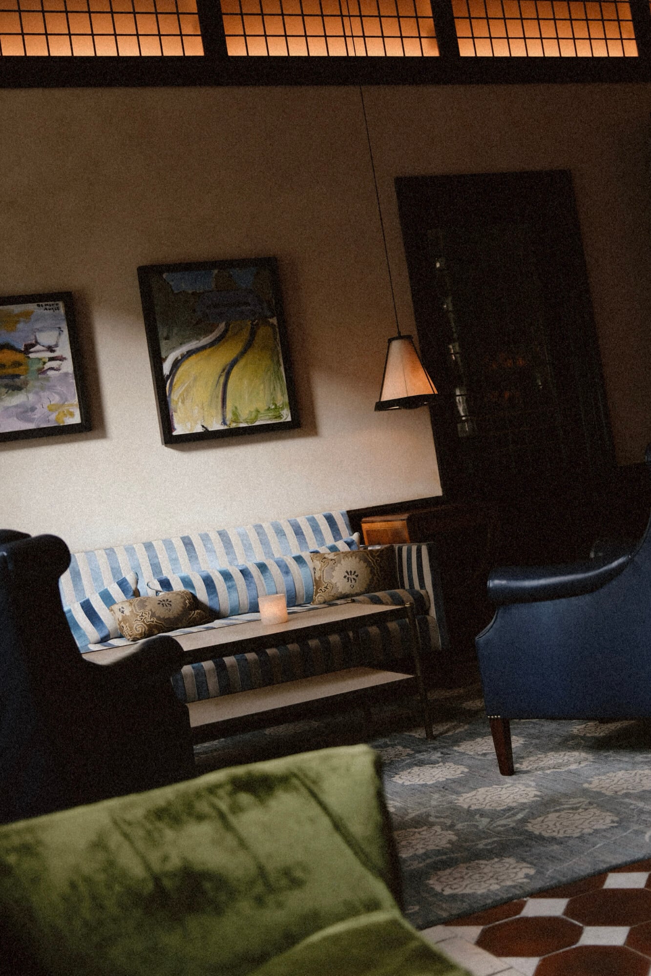 The Greenwich Hotel, North America — Mid-century modern lounge with striped sofa, leather chairs, pendant lighting, framed artwork, and skylight creating warm ambient atmosphere.