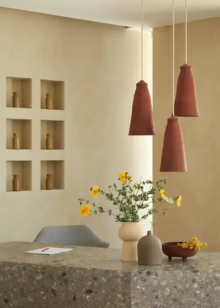 Pārocks, Europe — Terrazzo table with pendant lamps, yellow flowers, and built-in shelving in minimalist hospitality interior.