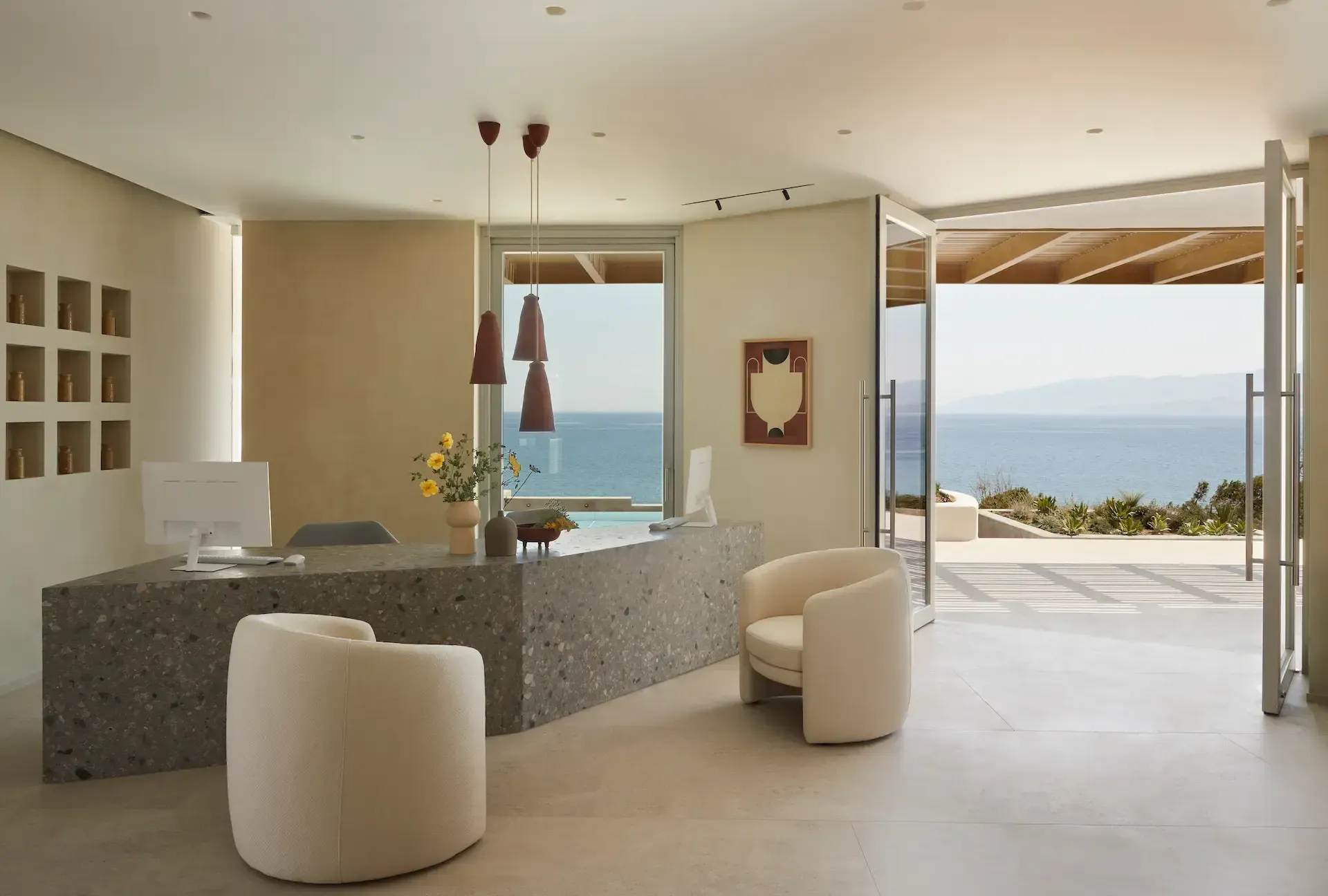 Pārocks, Europe — Minimalist coastal reception with terrazzo desk, cream curved seating, pendant lights, and expansive ocean views through floor-to-ceiling windows.