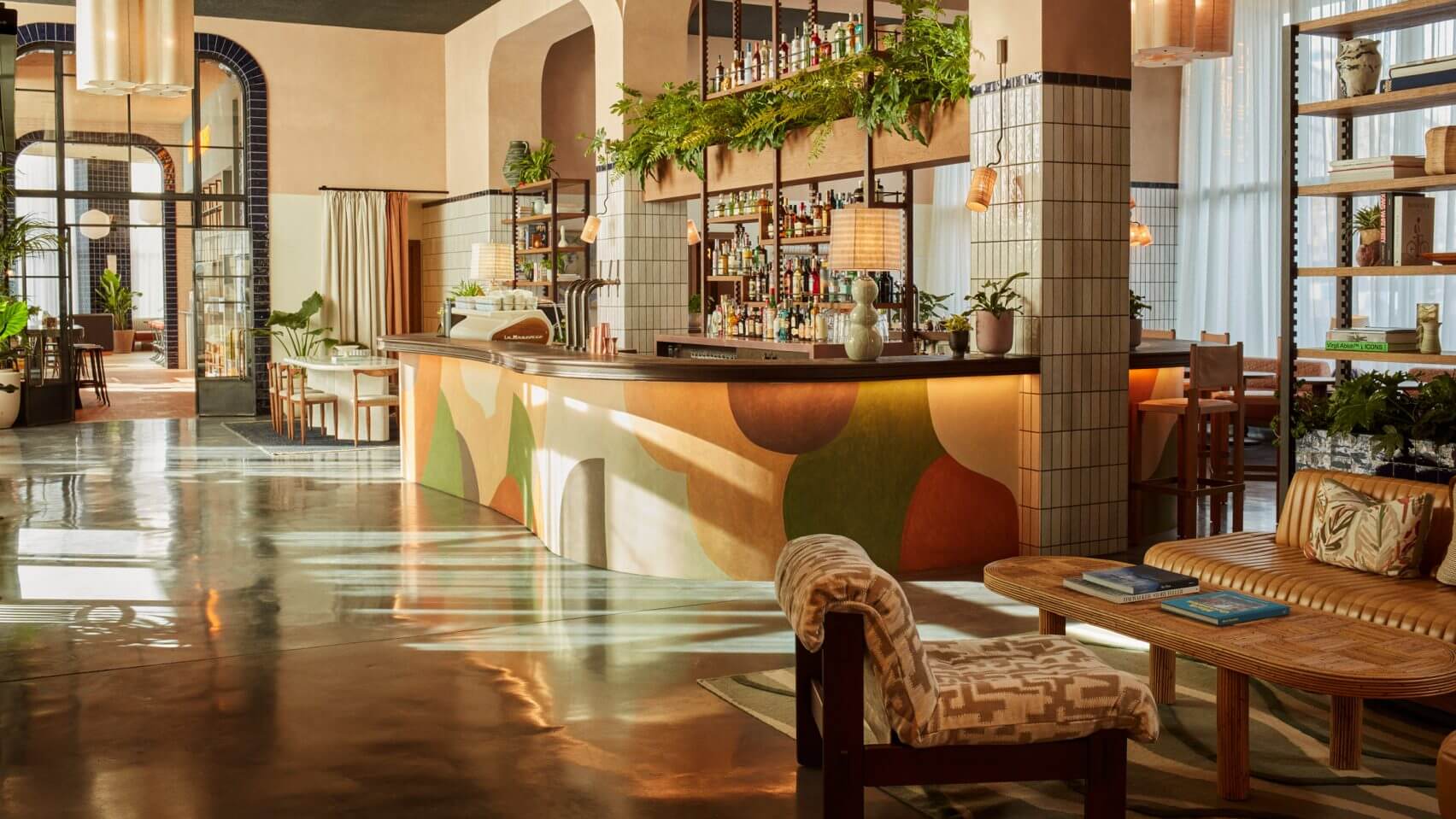 The Hoxton Poblenou, Barcelona, Spain — Sunlit hotel lobby with curved bar, geometric tile work, hanging plants, terracotta tones, and modernist furnishings.