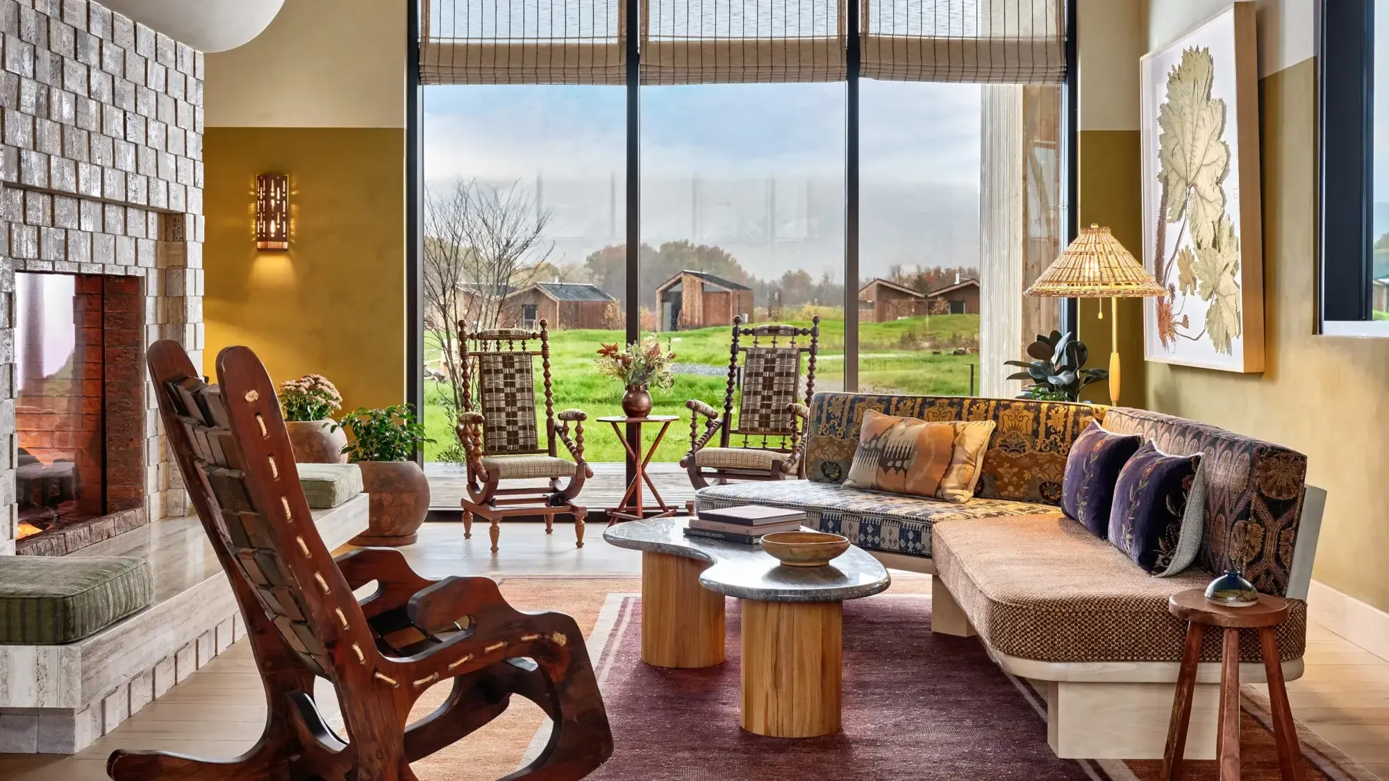 Wildflower Farms, North America — Sunlit living room with mid-century modern seating, warm ochre walls, rural landscape views, and wooden accent tables.
