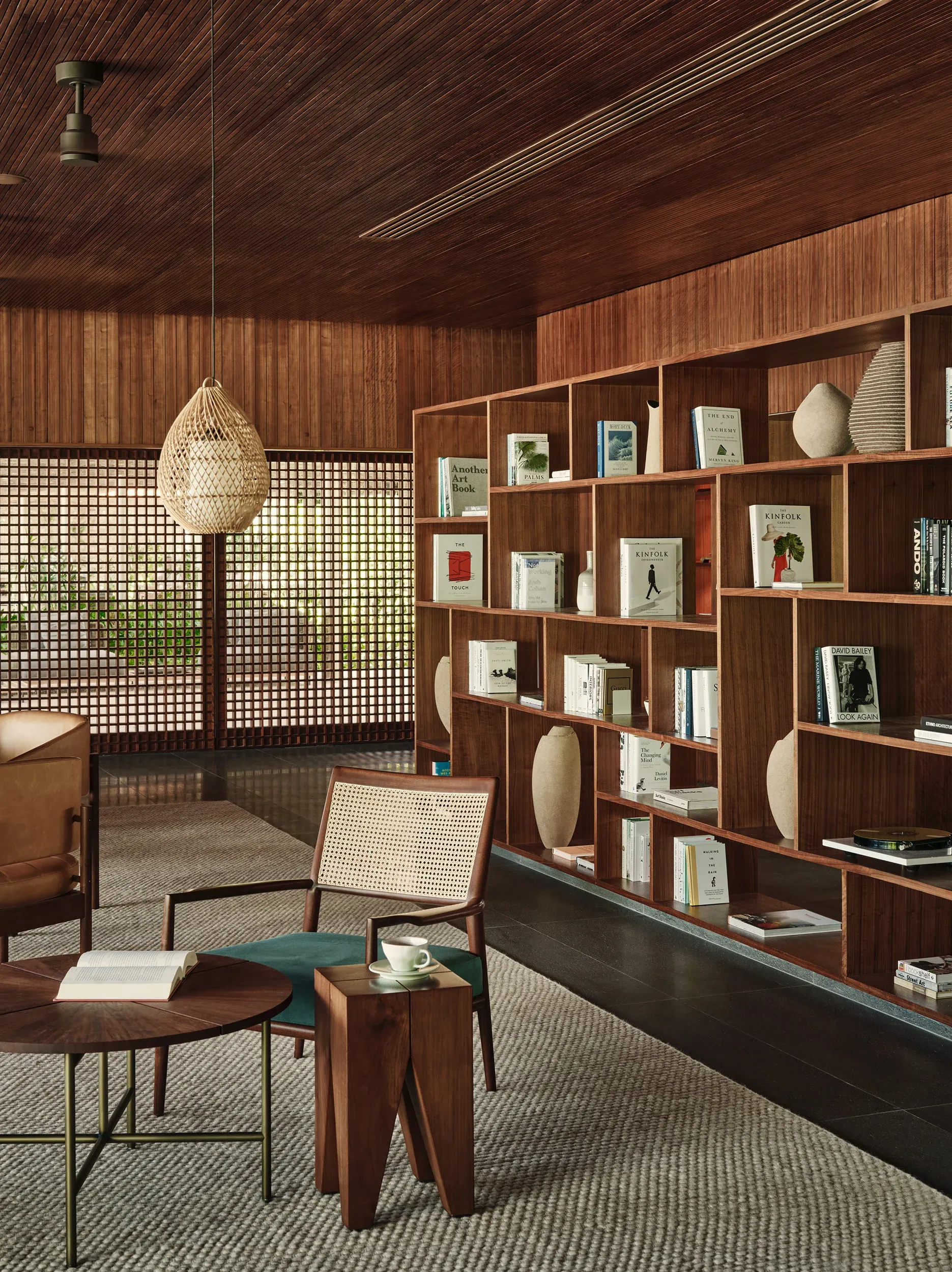 Patina Maldives, Asia — Wooden library lounge with woven pendant light, mid-century chair, and floor-to-ceiling shelving displaying books and sculptural objects.