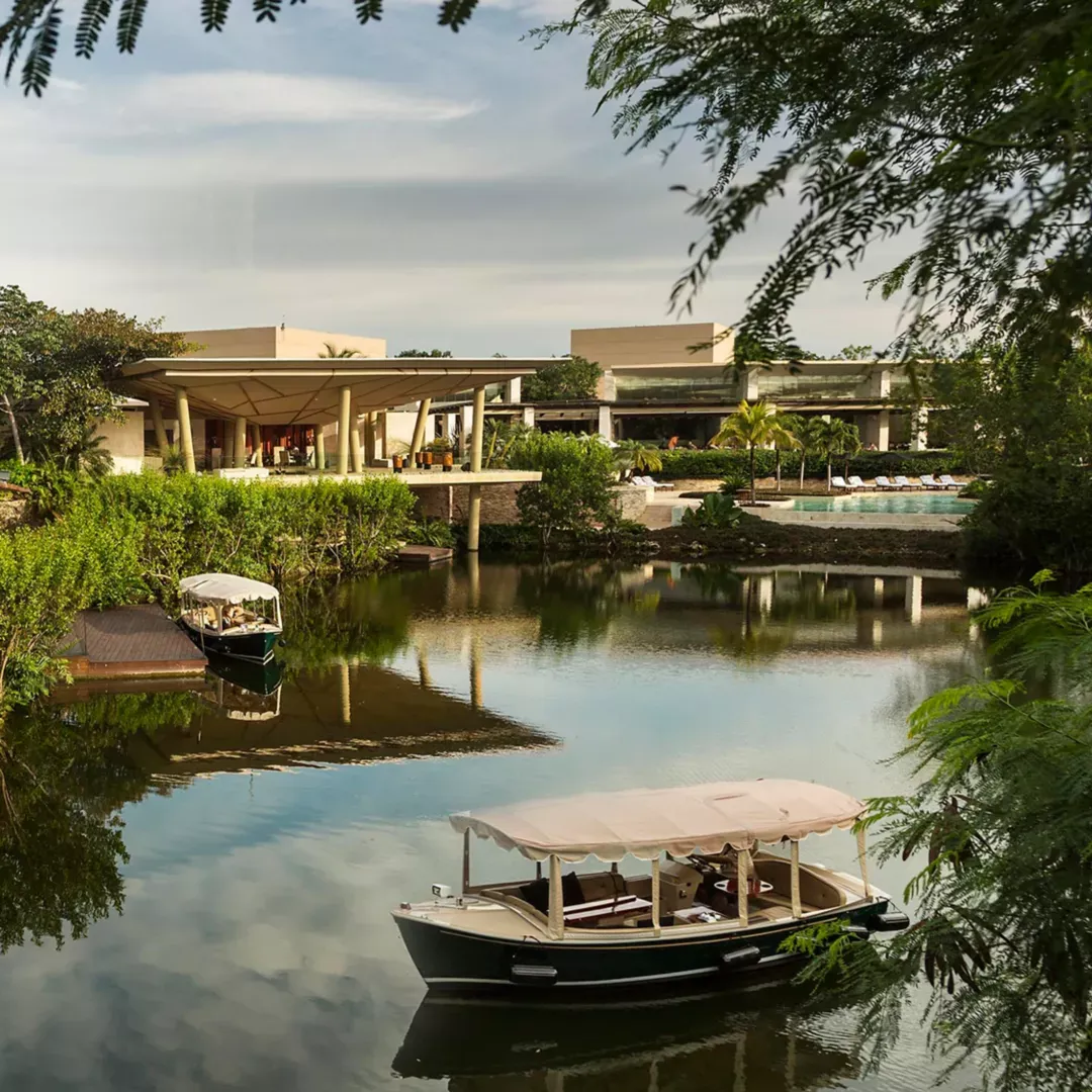 Rosewood Mayakoba, Mexico — Modernist waterfront pavilion with limestone columns overlooking mangrove lagoon, traditional boat moored at dock, tropical vegetation, evening light.