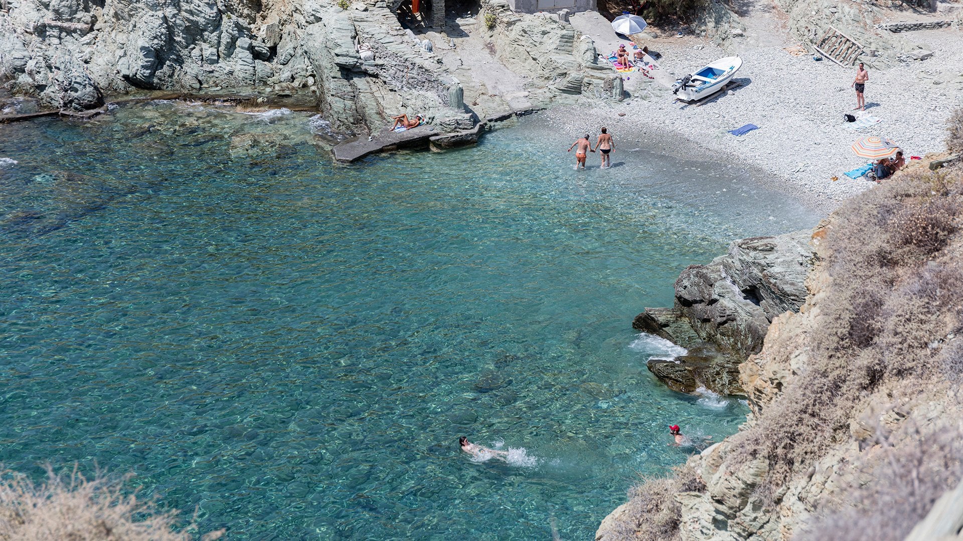 Local Beaches, Greece — Turquoise cove with limestone cliffs, moored boats, sunbathers on pebble beach, crystal-clear Mediterranean waters.