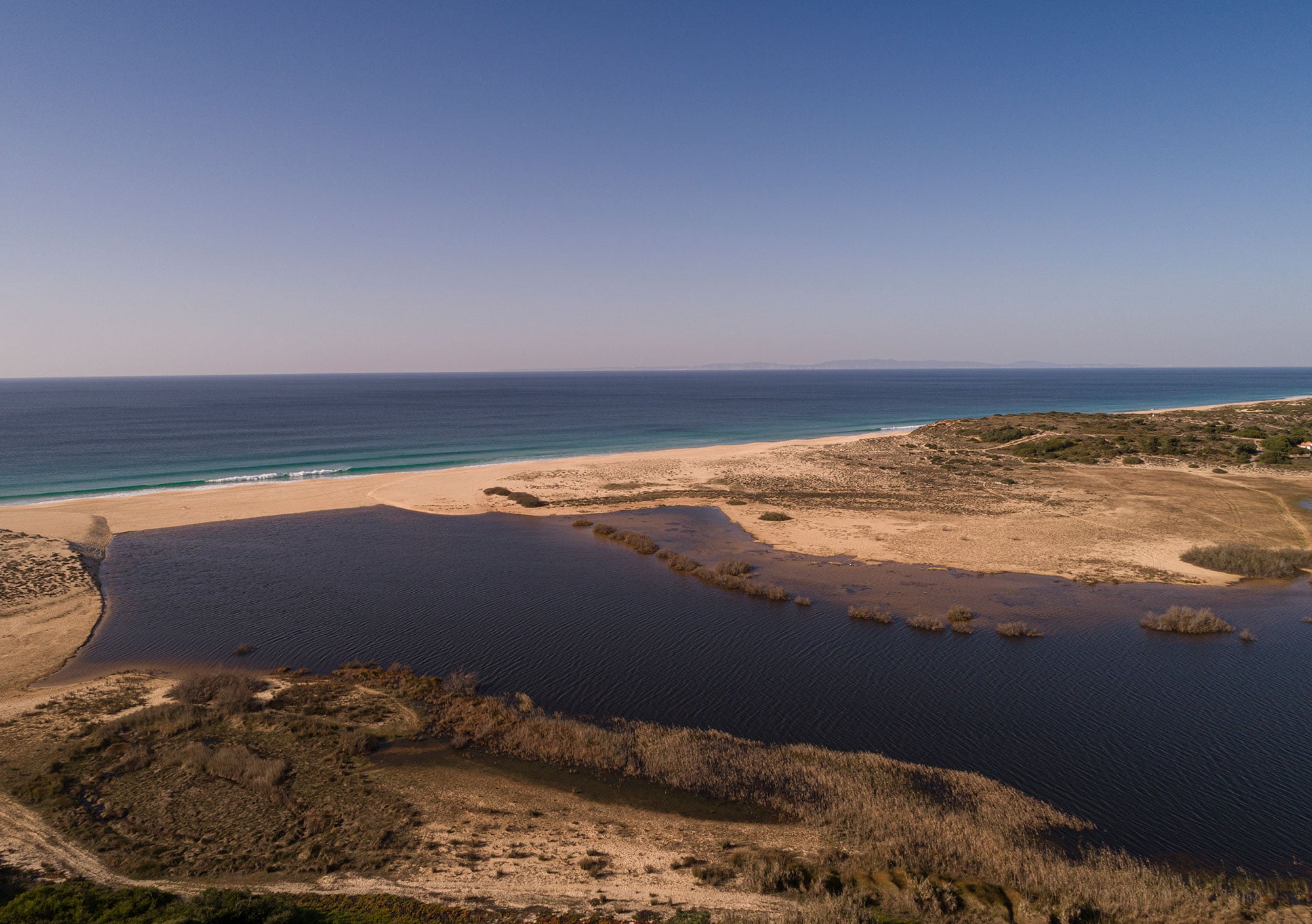 Aerial view of Praia de Melides lagoon with dark brackish water, golden sand beach, and turquoise ocean under clear sky.