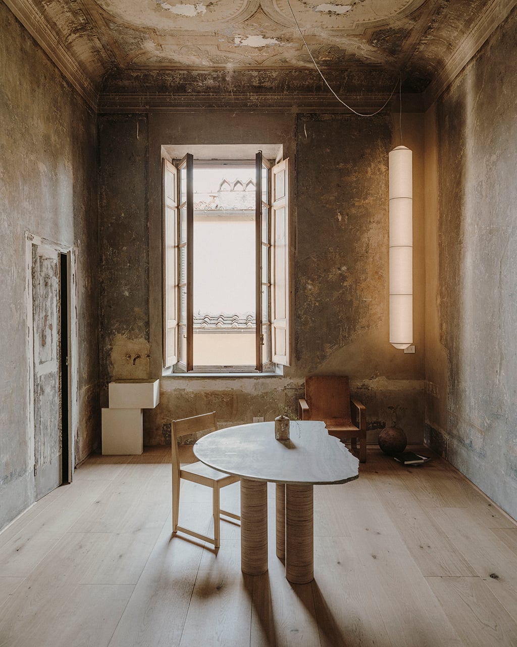 Numeroventi, Europe — Minimalist room with distressed plaster walls, pale wood flooring, oval marble table, and tall cylindrical pendant light framing tall windows overlooking roofscape.
