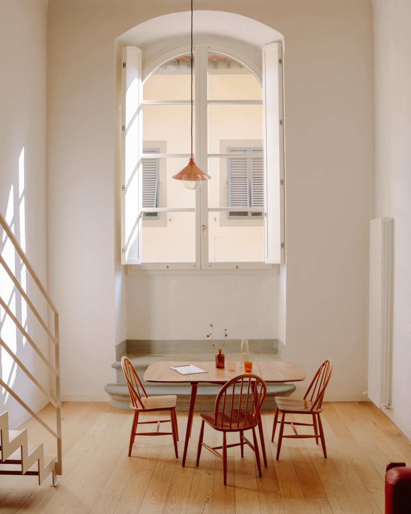 Numeroventi, Europe — Minimalist dining room with arched window, copper pendant light, wooden chairs, and natural stone countertop overlooking courtyard.