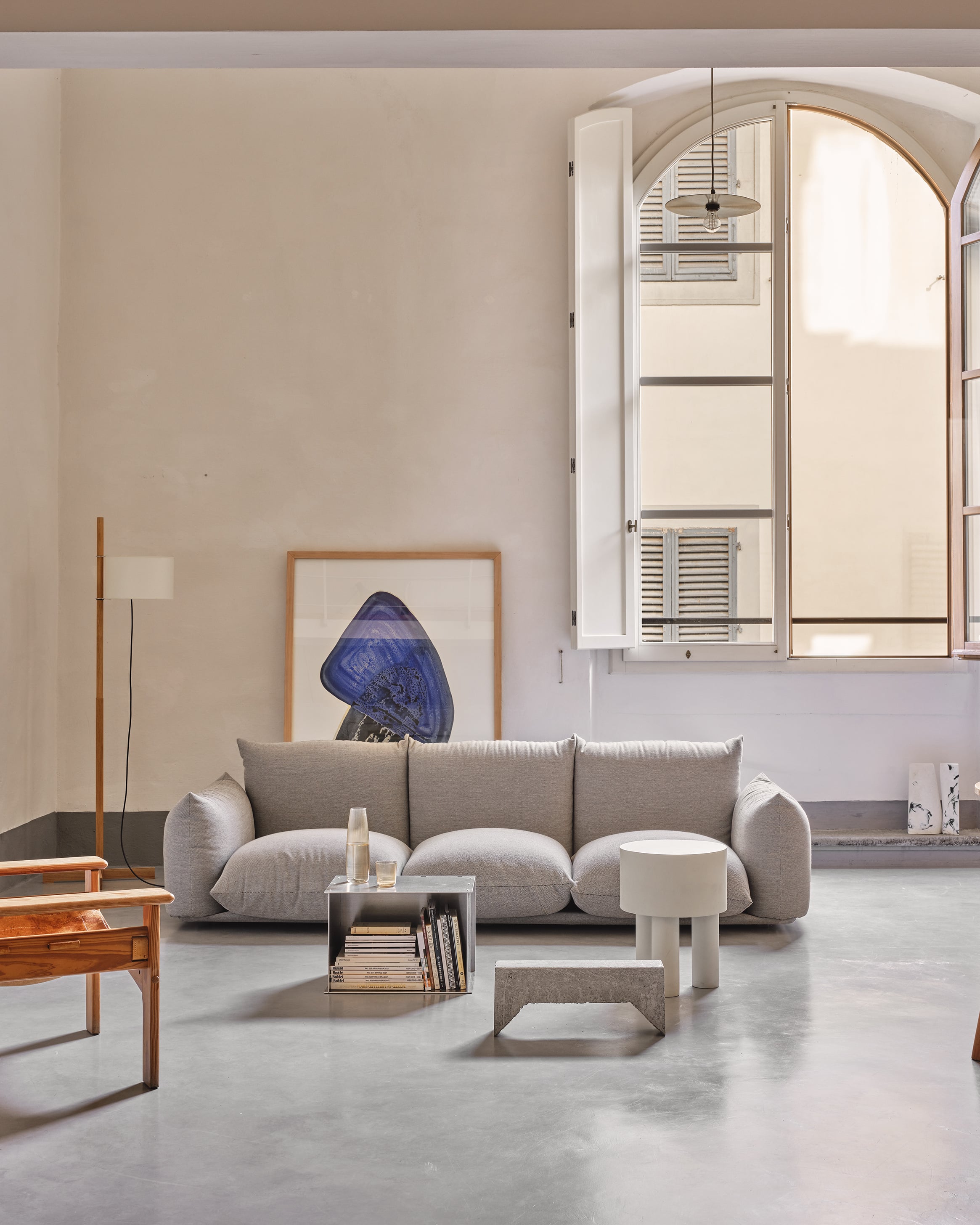 Numeroventi, Europe — Minimalist living room with curved gray sofa, arched windows, concrete floors, wooden furniture, and blue artwork under natural light.