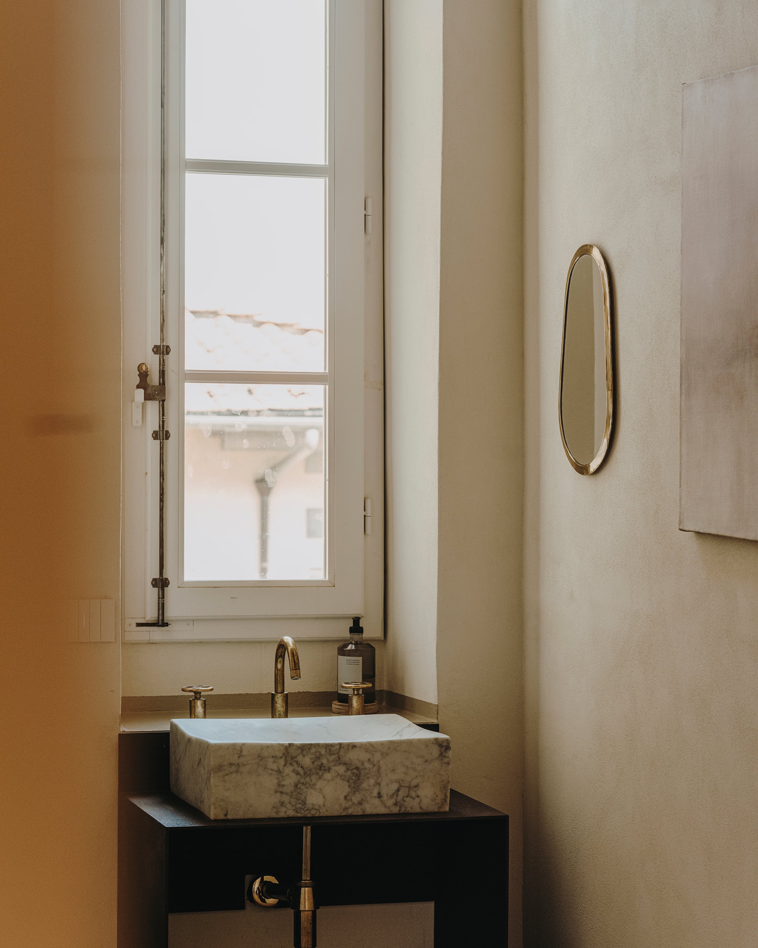 Numeroventi, Europe — Minimalist bathroom with marble vessel sink on black vanity, brass fixtures, oval mirror, and tall window overlooking roofscape.