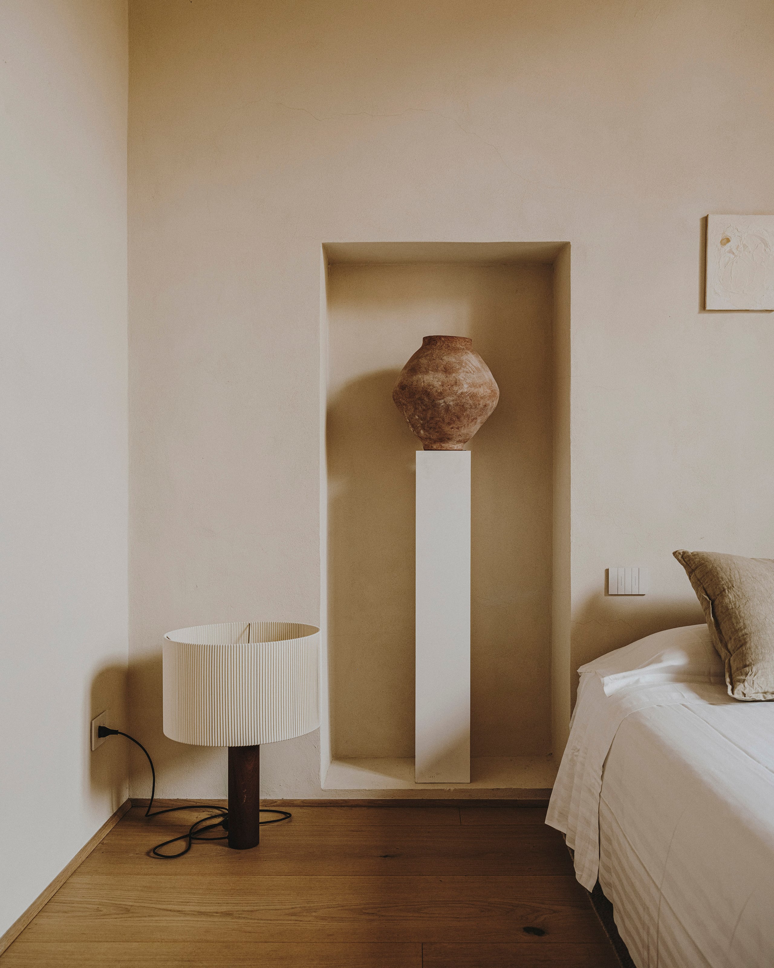 Numeroventi, Europe — Minimalist bedroom with recessed wall niche displaying terracotta vessel on pedestal, ribbed table lamp, wooden flooring, neutral palette.