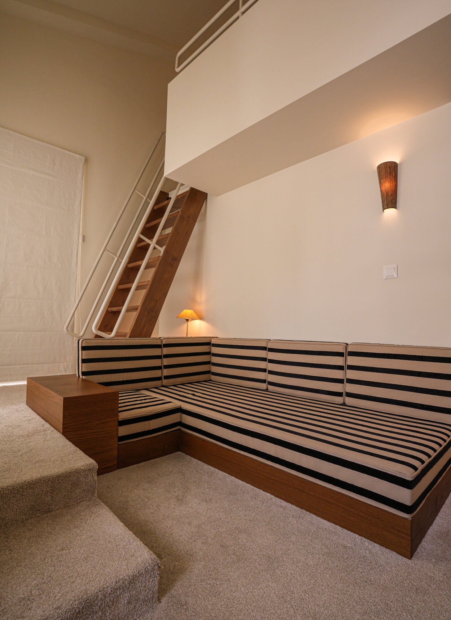Apollo Palm, Europe — Minimalist hotel bedroom with striped black and white bedding, wooden platform bed, concrete ceiling, warm wall sconces, and architectural staircase detail.