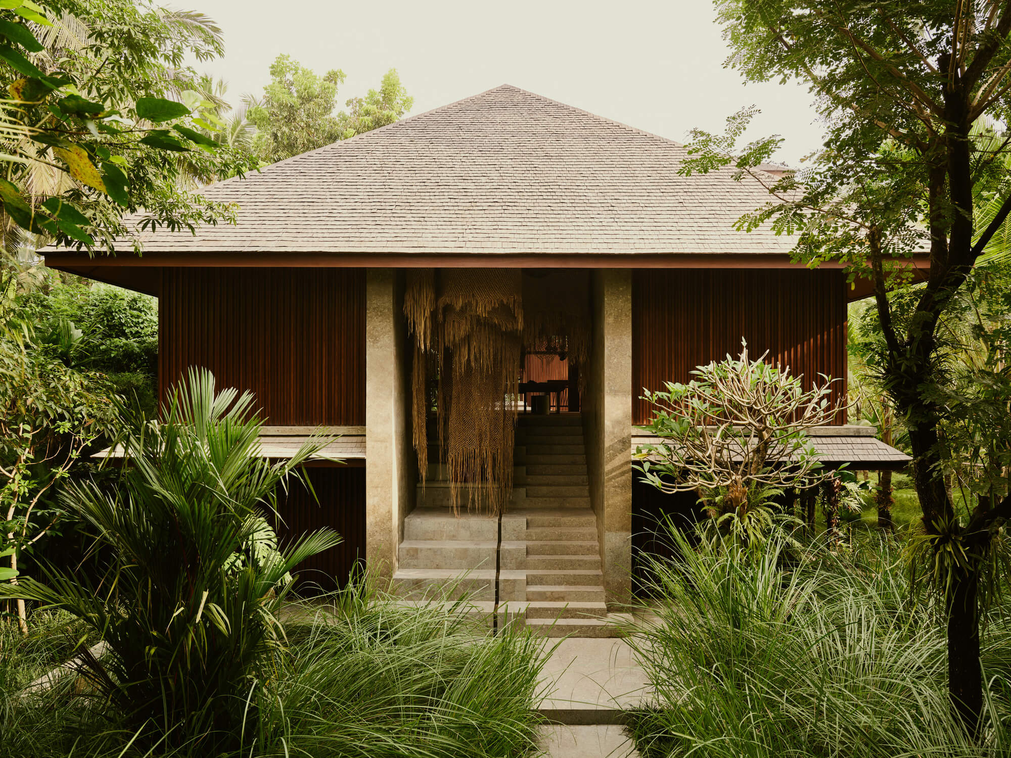 Lost Lindenberg, Asia — Concrete and timber pavilion with pyramid roof nestled in lush tropical gardens, stone steps leading to open interior with woven ceiling detail.