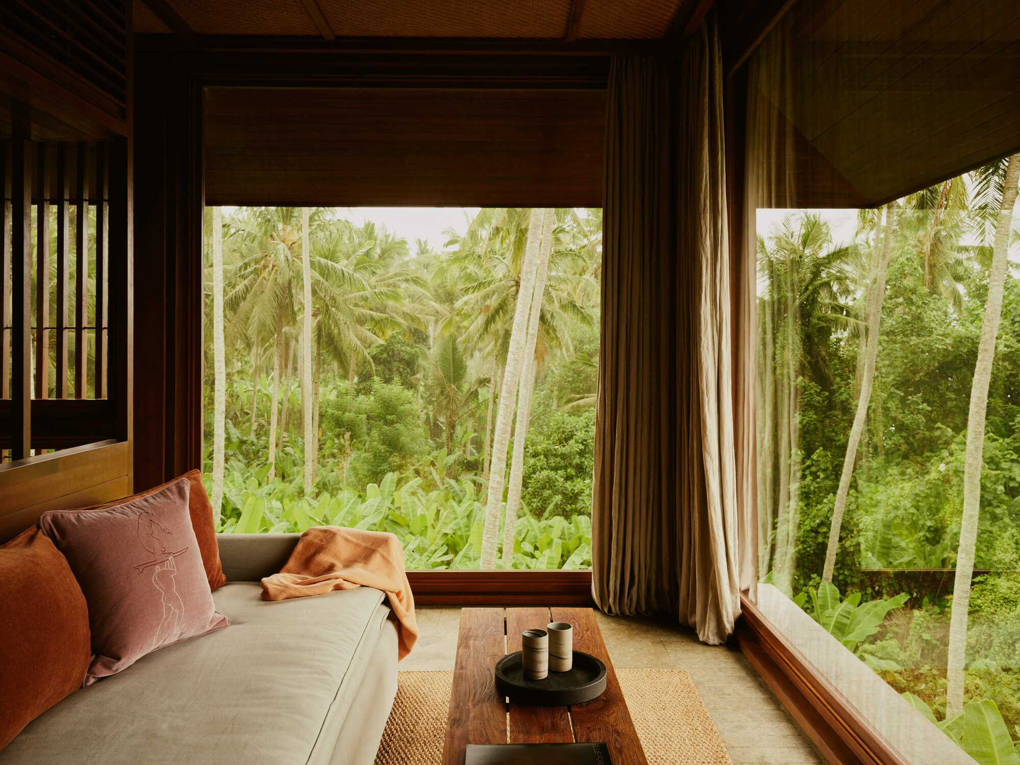 Lost Lindenberg, Asia — Bedroom pavilion with floor-to-ceiling windows framing tropical forest canopy, teak furnishings, linen upholstery, and natural light filtering through floor-to-ceiling curtains.