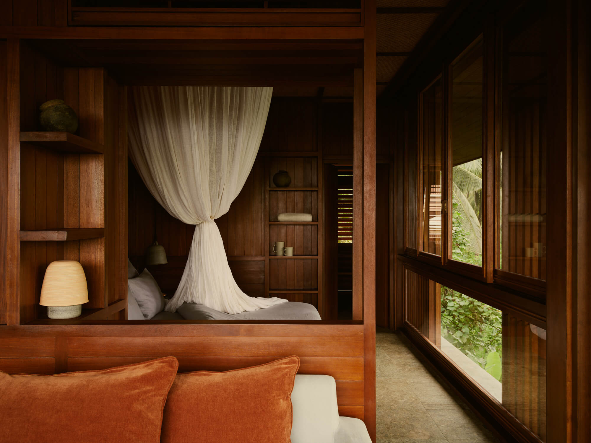 Lost Lindenberg, Asia — Wooden canopy bed with sheer white curtains in dark timber bedroom overlooking lush tropical garden through louvered windows.
