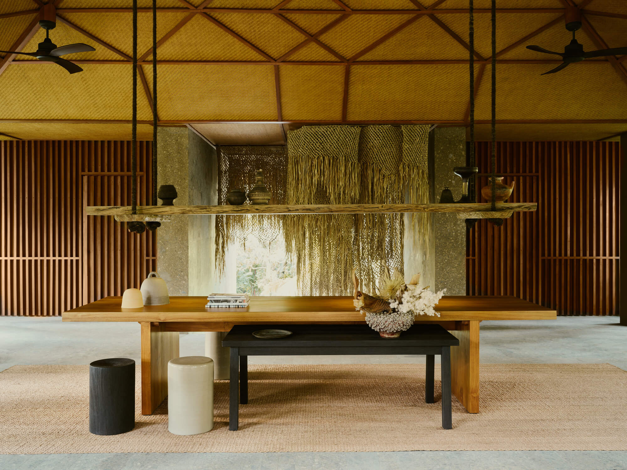 Lost Lindenberg, Asia — Minimalist lounge with timber table, woven macramé wall hanging, corrugated metal ceiling, and natural light filtering through layered textiles.