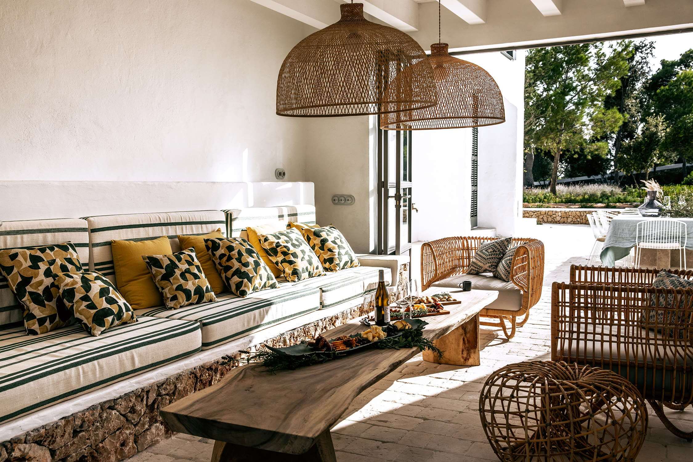 Es Bec d’Aguila — Es Bec d'Aguila, Balearic Islands, Spain — Covered terrace with built-in stone seating, geometric patterned pillows, woven pendant lights, wooden furniture, and garden views.