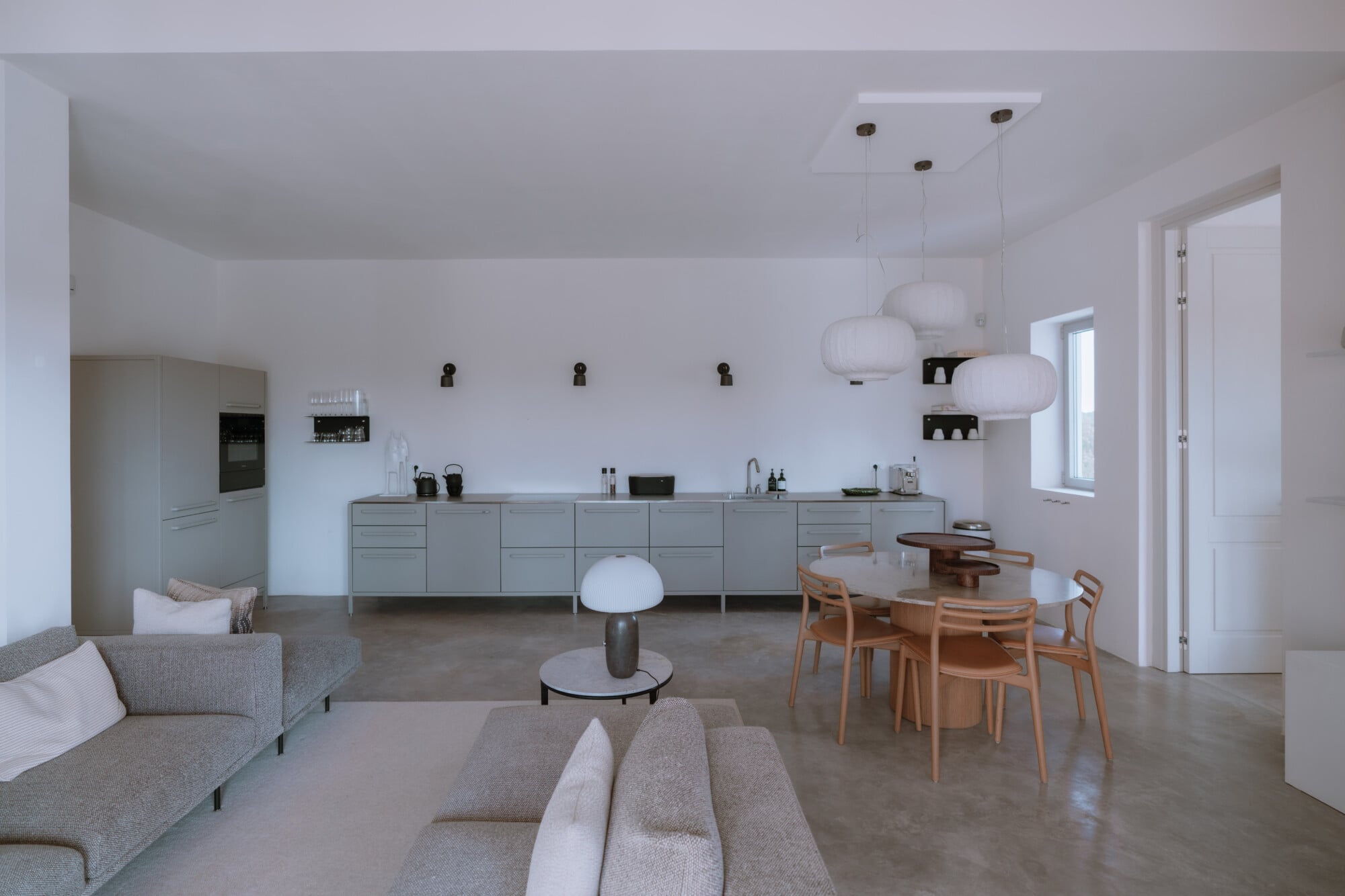 Villa Quercia, Italy — Minimalist open-plan living space with pale grey kitchen cabinetry, natural wood dining chairs, linen sofas, pendant lighting, and polished concrete flooring.