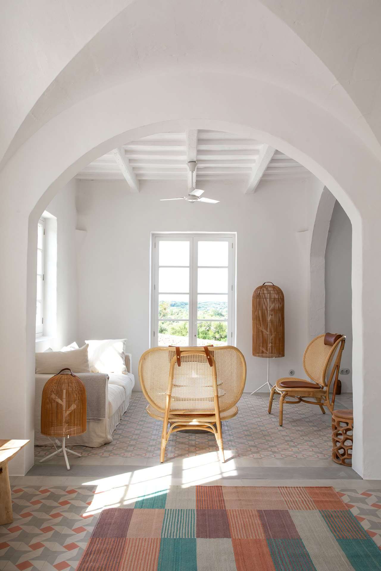 Es Bec d’Aguila — Es Bec d'Aguila, Balearic Islands, Spain — Arched white room with rattan chairs, timber ceiling, panoramic windows overlooking landscape, terracotta and geometric floor tiles.
