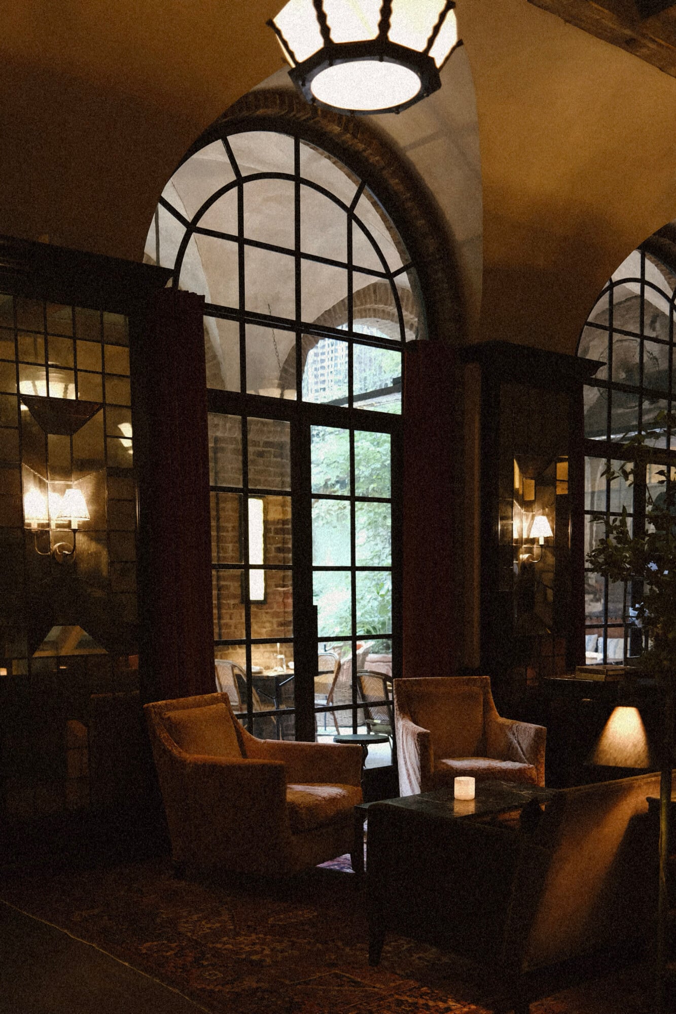 Drawing Room & Courtyard, North America — Arched windows with grid pattern frame courtyard view, warm lighting on ochre walls, leather chairs, minimalist luxury hospitality interior.