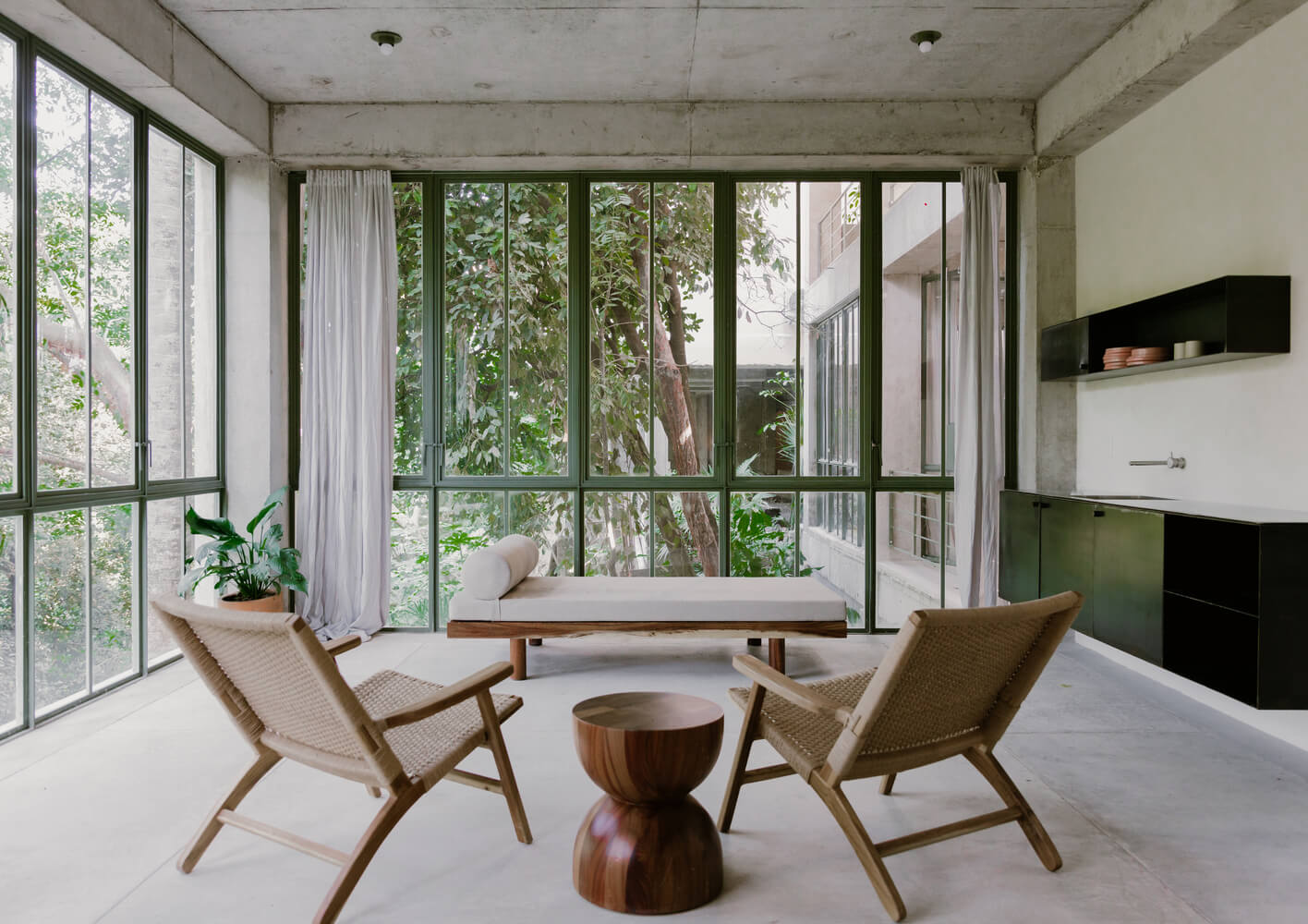 NICO, Mexico — Modernist bedroom with floor-to-ceiling glazing, concrete ceiling, woven lounge chairs, wooden side table, and verdant garden views.