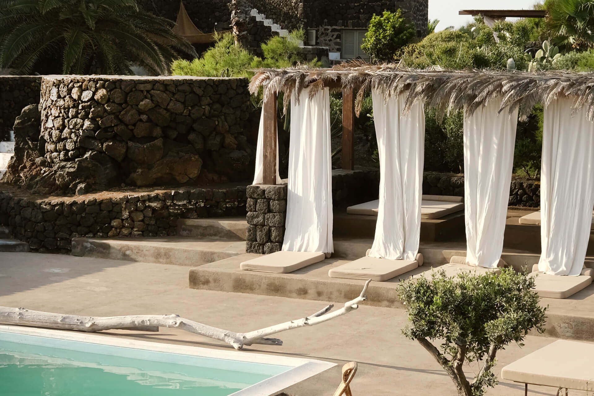Parco Dei Sesi, Europe — Poolside pergola with flowing white curtains, stone walls, and Mediterranean garden landscape in warm sunlight.