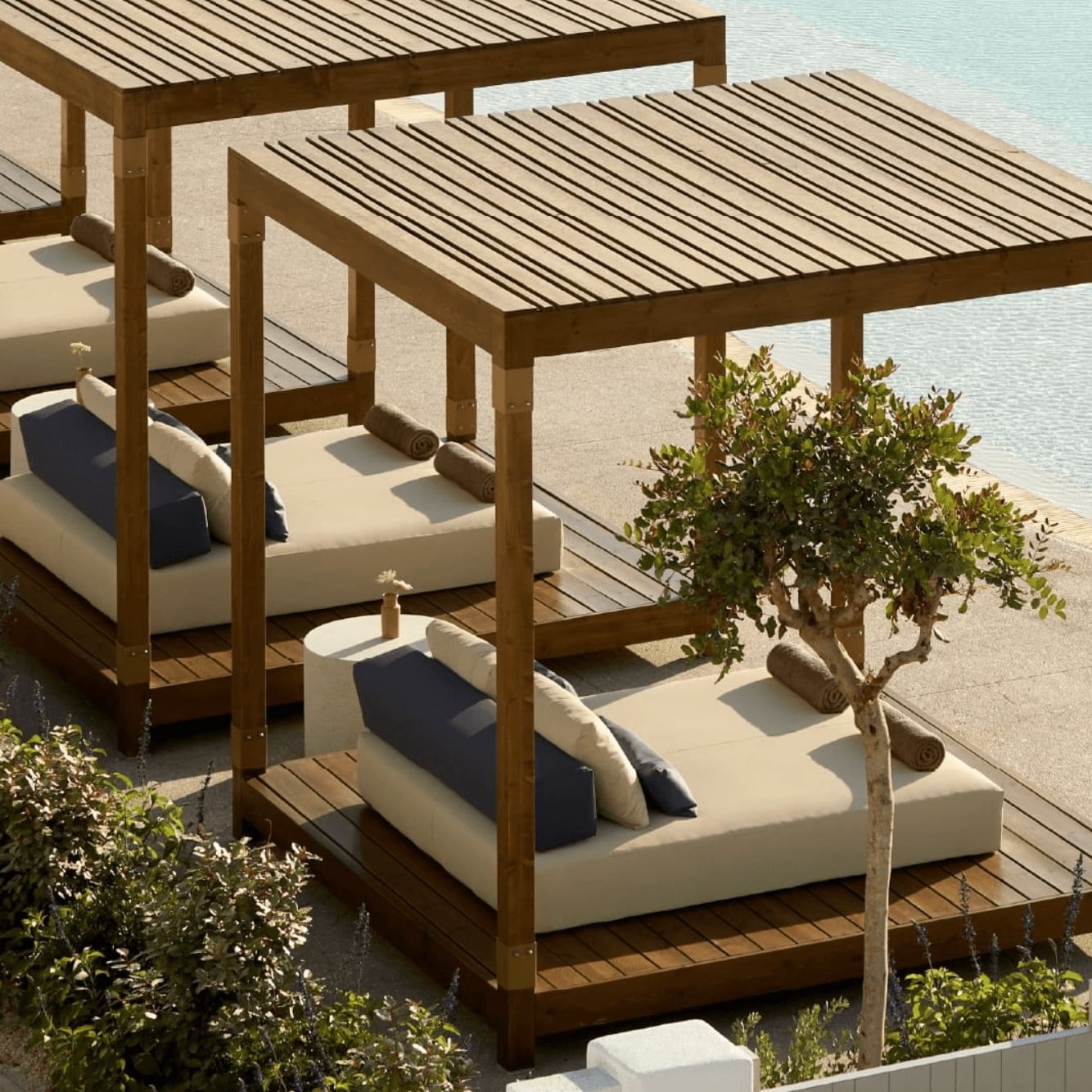 Pārocks, Europe — Wooden pergola cabanas with cream loungers and navy pillows overlook water, framed by olive trees and warm natural light.