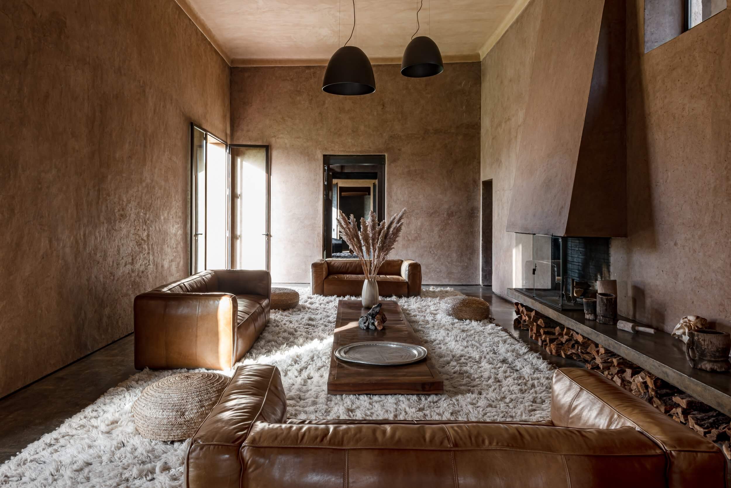 Villa D, Africa — Minimalist living room with cognac leather seating, cream shag rugs, textured terracotta walls, and linear fireplace beneath black pendant lights.