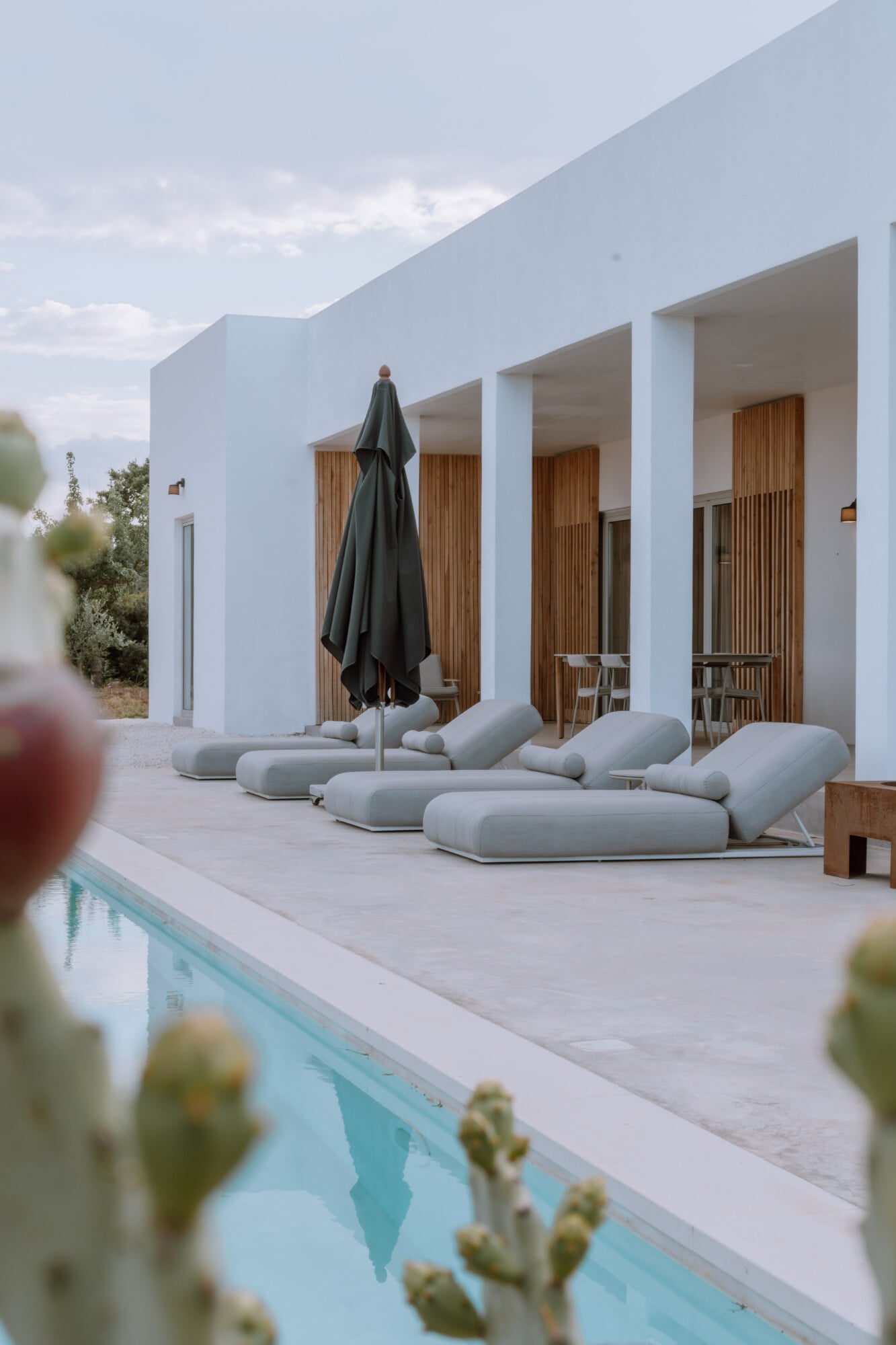 Villa Quercia, Italy — Modernist white villa with wooden-shuttered openings, gray loungers, black umbrella, and swimming pool edge with cacti foreground.
