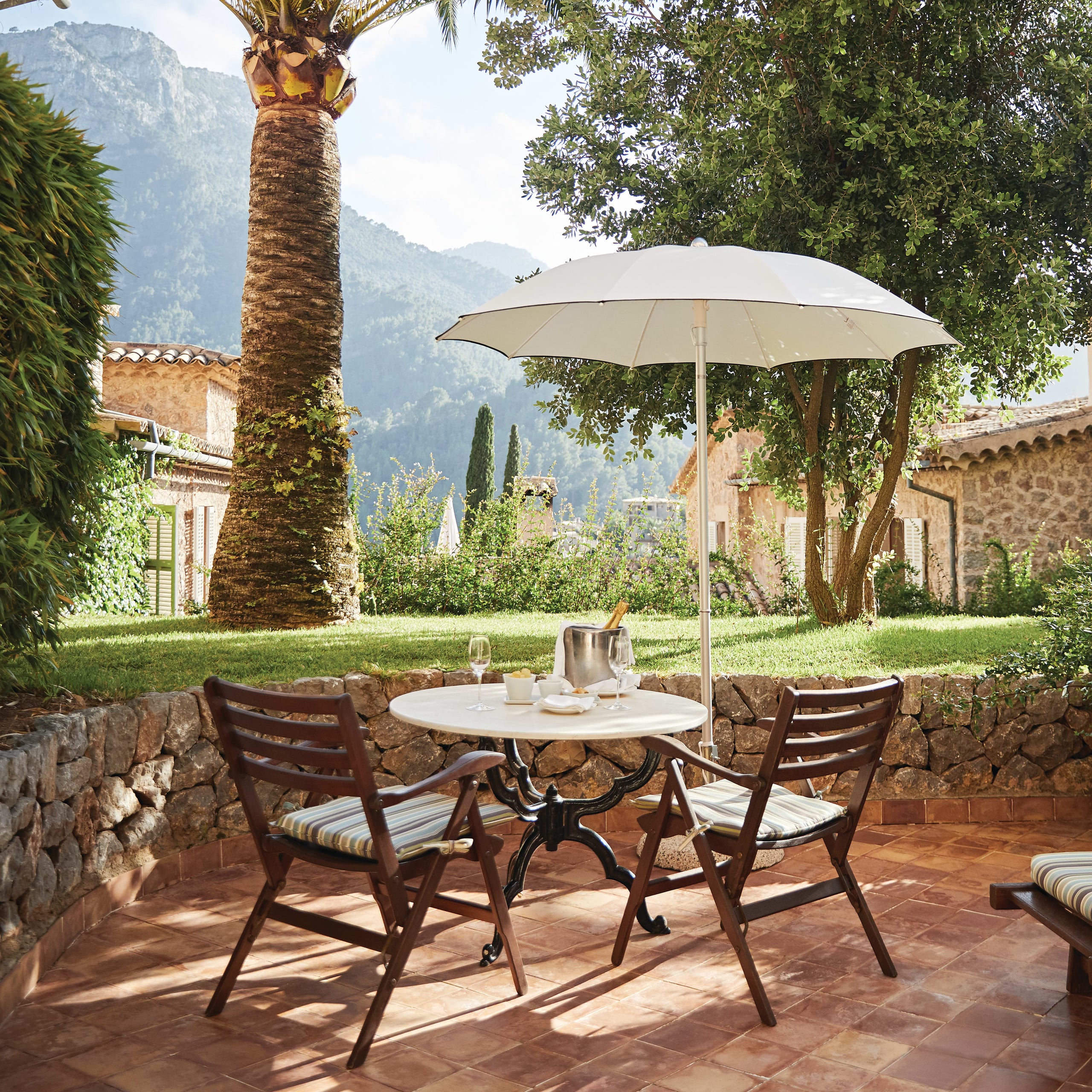 Belmond La Residencia, Balearic Islands, Spain — Shaded terracotta patio with round table, wooden chairs, umbrella, stone wall, palm tree, and mountain views across manicured gardens.