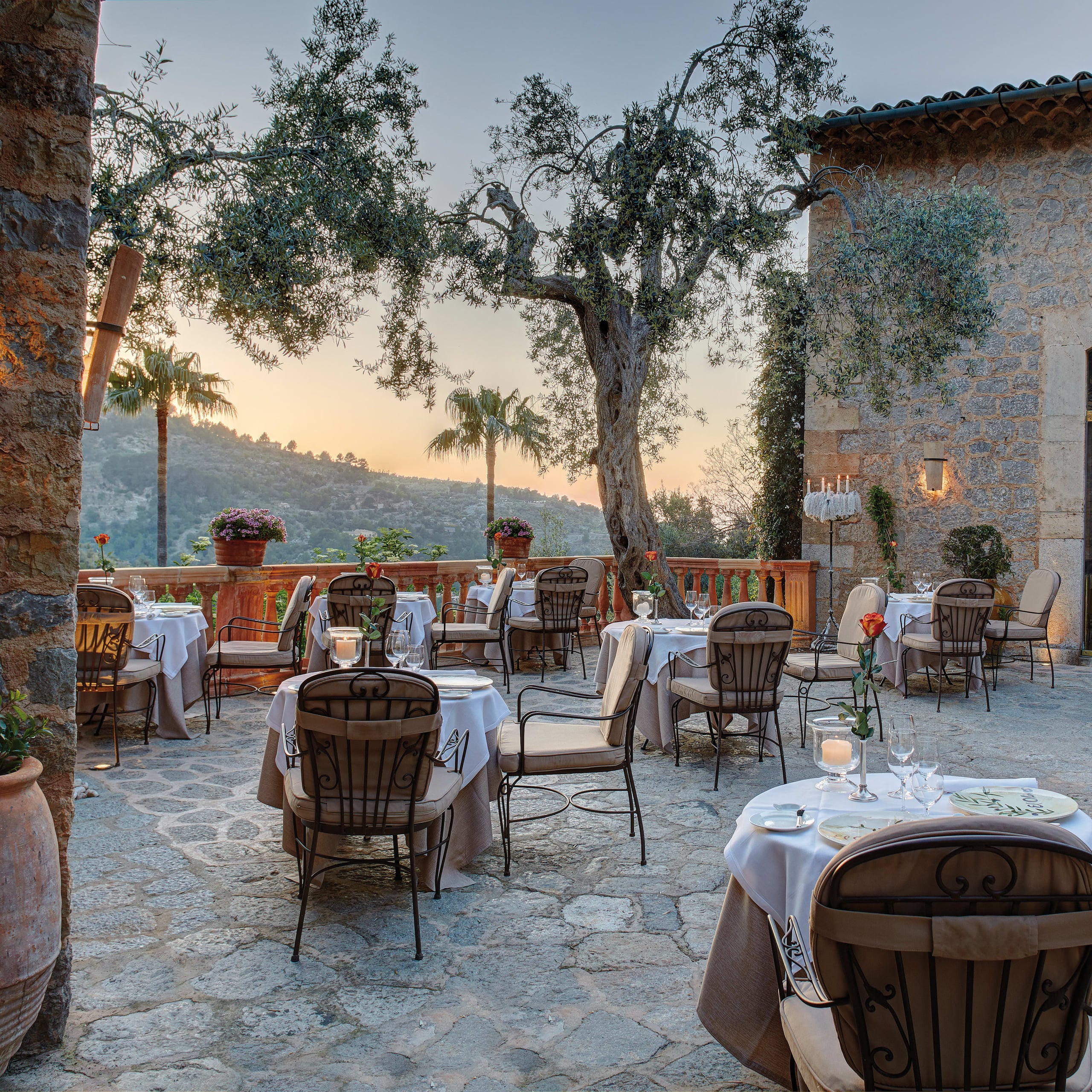 Belmond La Residencia, Balearic Islands, Spain — Candlelit dining terrace at dusk with wrought-iron chairs, white linens, ancient olive tree, stone architecture, and mountainous landscape beyond.