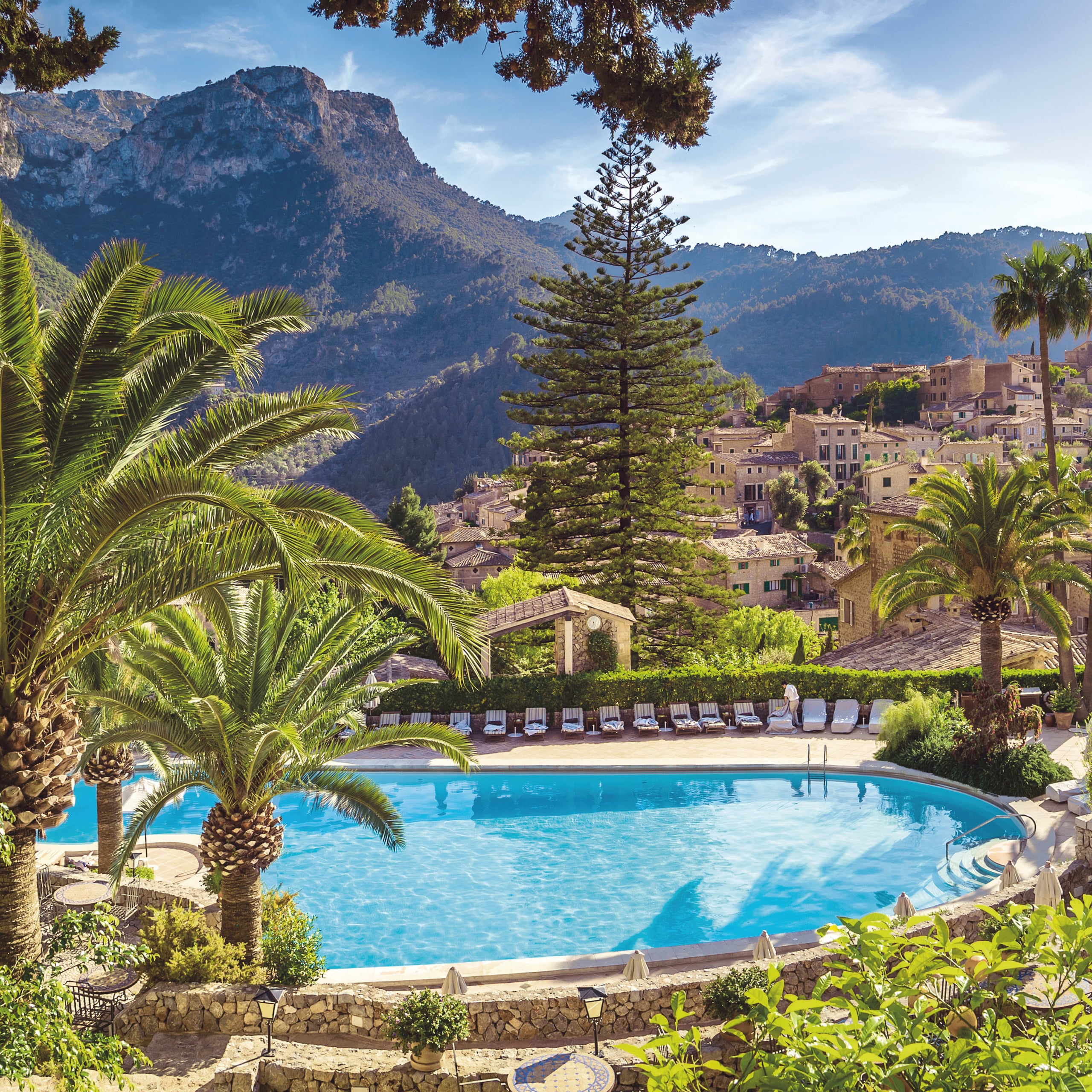 Belmond La Residencia, Balearic Islands — Resort pool framed by palm trees with stone terrace, mountainous backdrop, and traditional Mediterranean architecture.