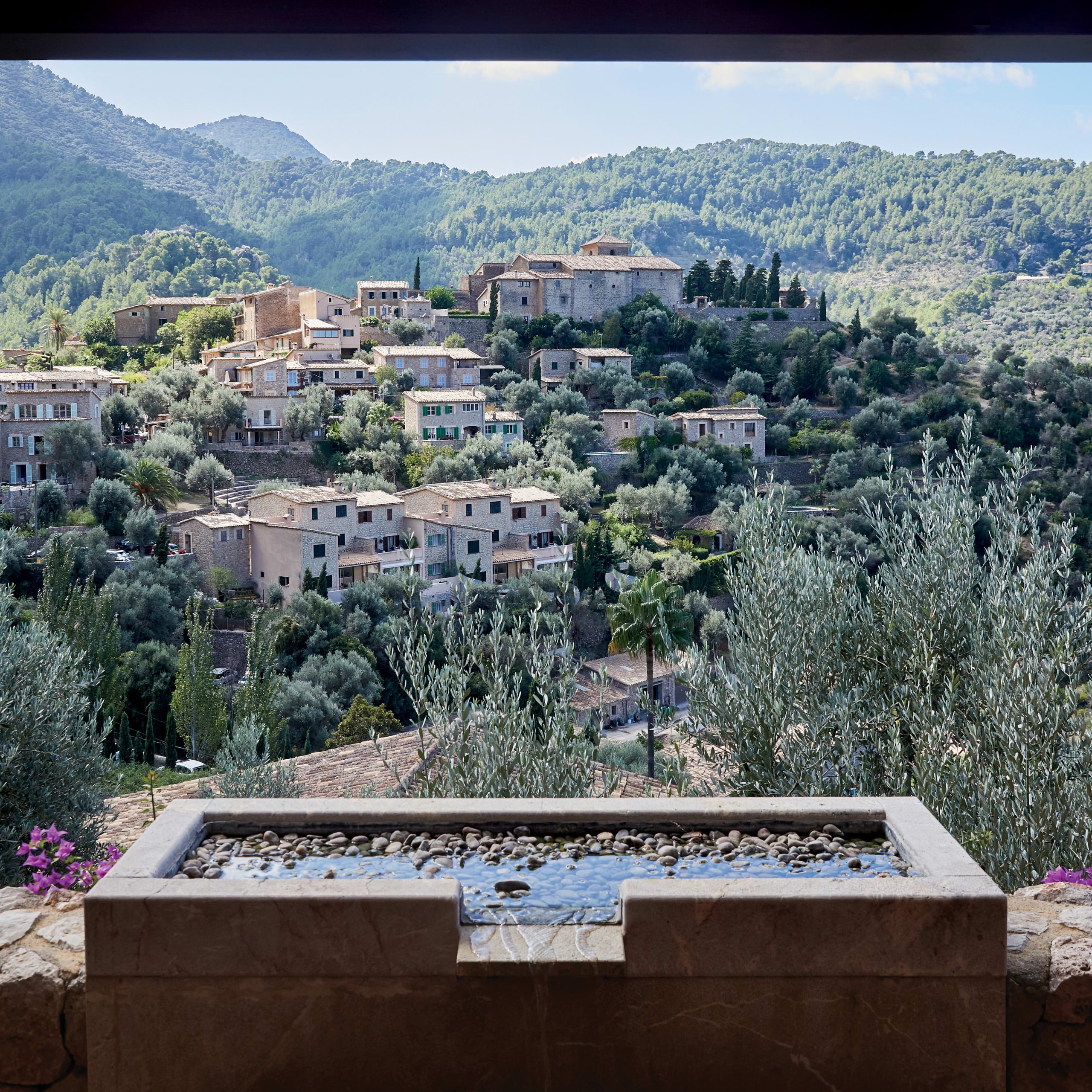 Belmond La Residencia, Balearic Islands — Terraced stone water feature overlooking Mediterranean village nestled in forested hills with traditional architecture.
